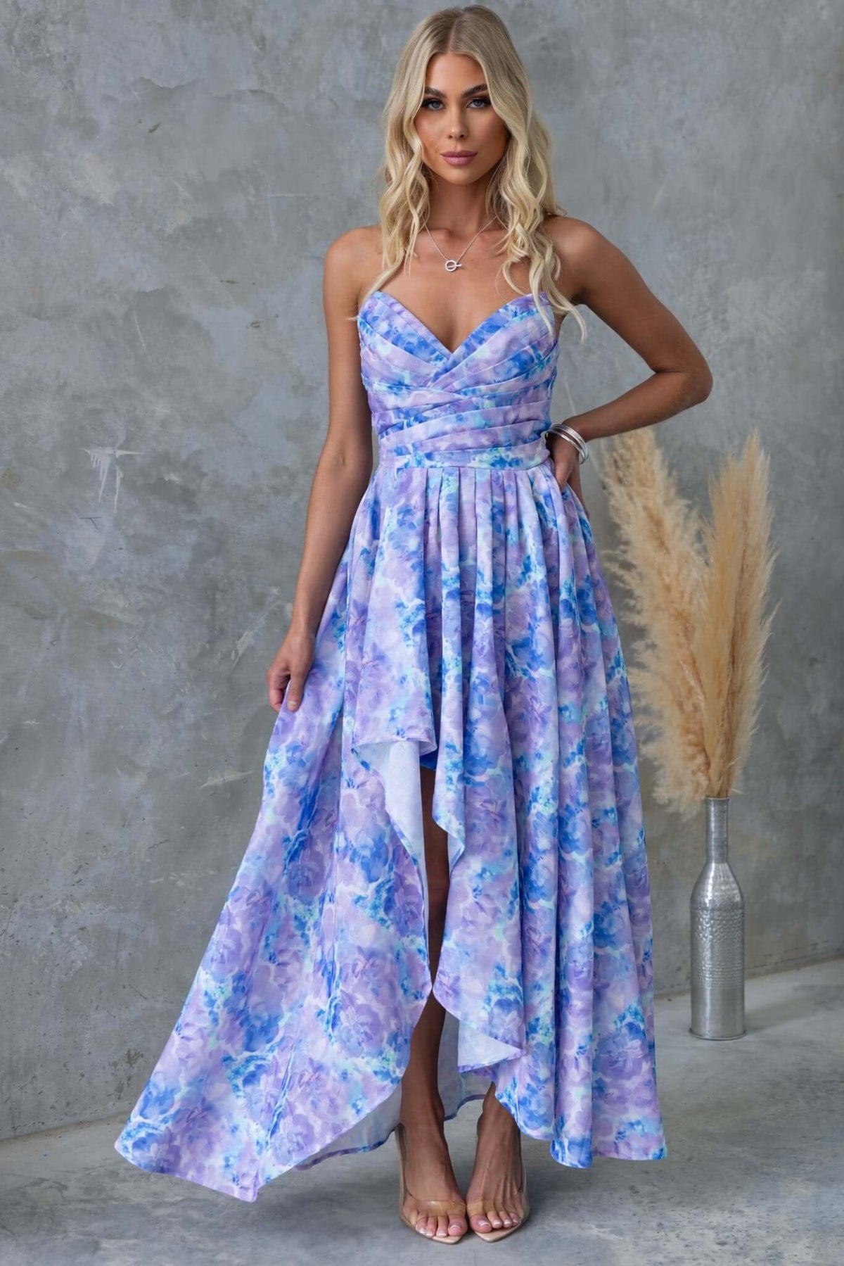 Model wearing Kiss And Tell Maxi Dress with strapless design, asymmetric hem, and floral pattern against a textured background.
