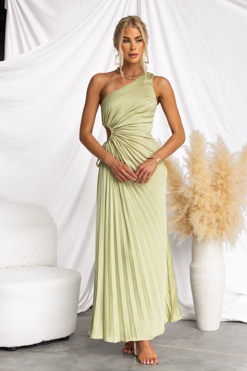 Model wearing a chic one-shoulder maxi dress in light green with pleats and a cut-out at the waist, styled with neutral decor.
