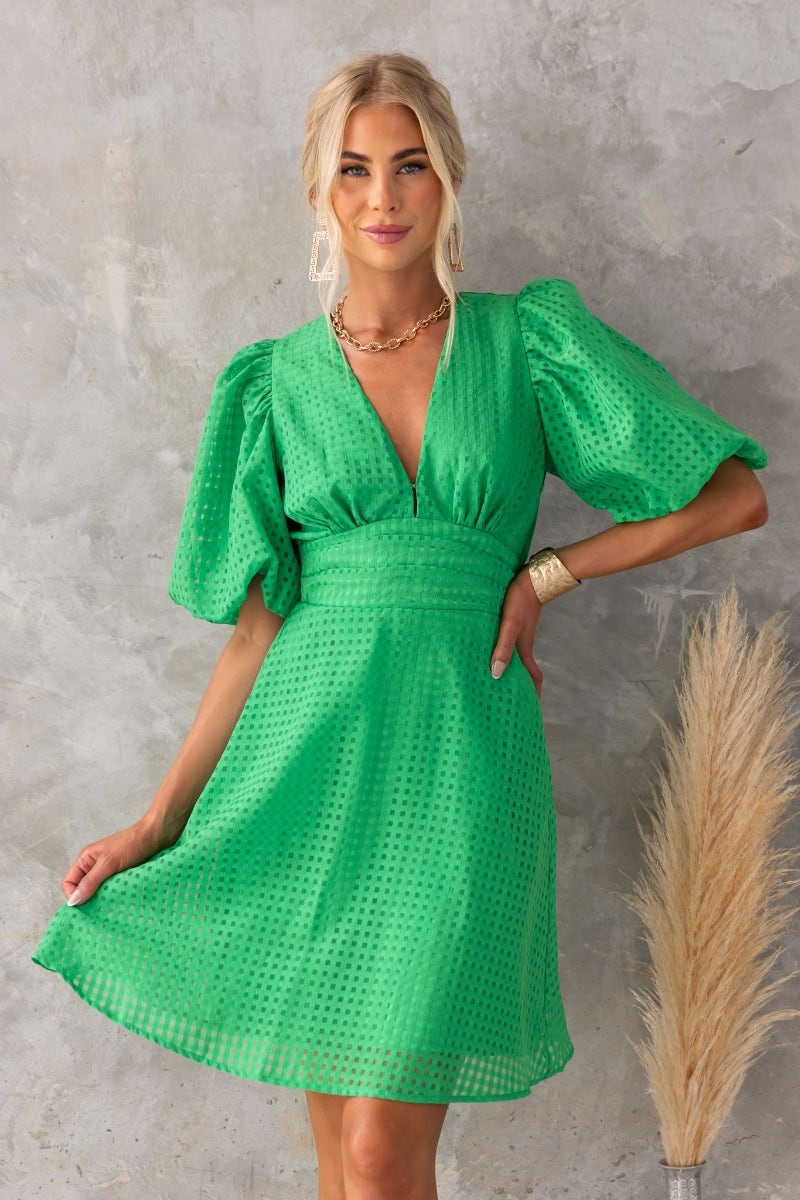 Lantier Dress in bright green with V-neckline, puff sleeves, and pockets, styled against a textured background.