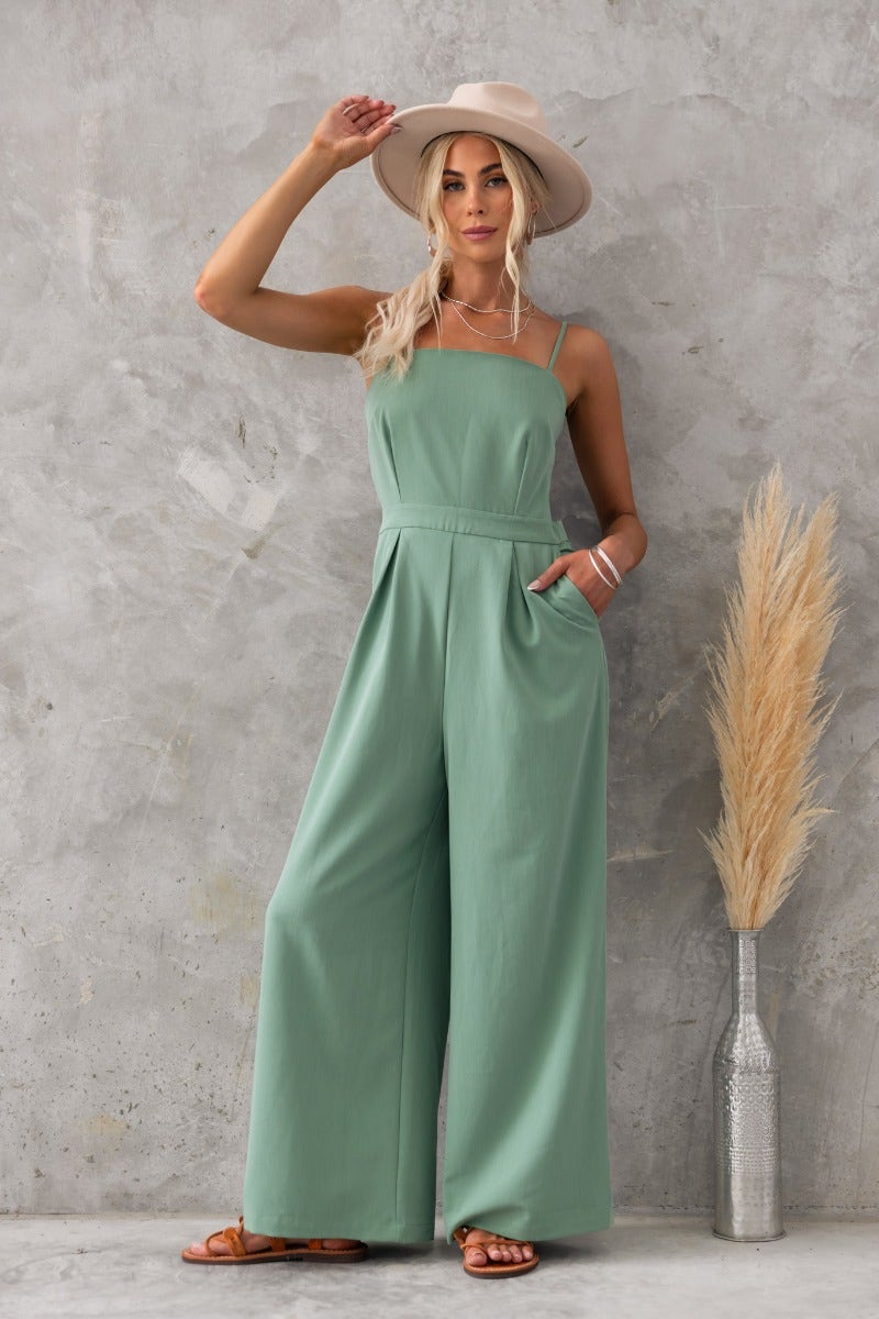 Model wearing Greta Jumpsuit in green with adjustable straps and pockets, styled with a hat and accessorized.