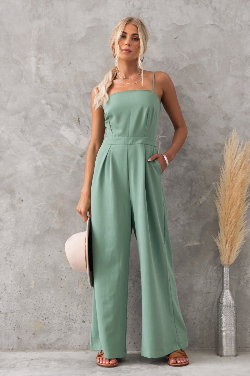 Model wearing a green Greta jumpsuit with adjustable straps, pockets, and a relaxed fit, set against a textured background.