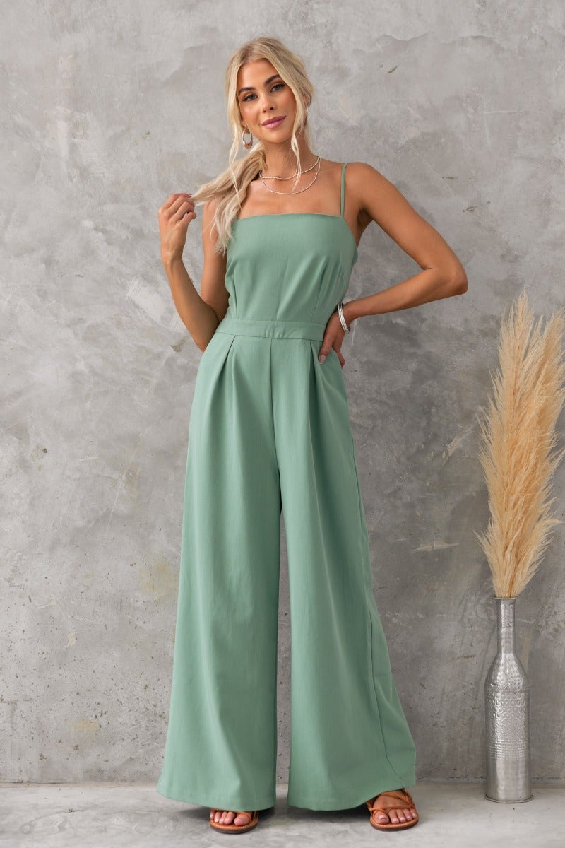 Model wearing a green Greta Jumpsuit with adjustable straps and wide legs against a textured backdrop.