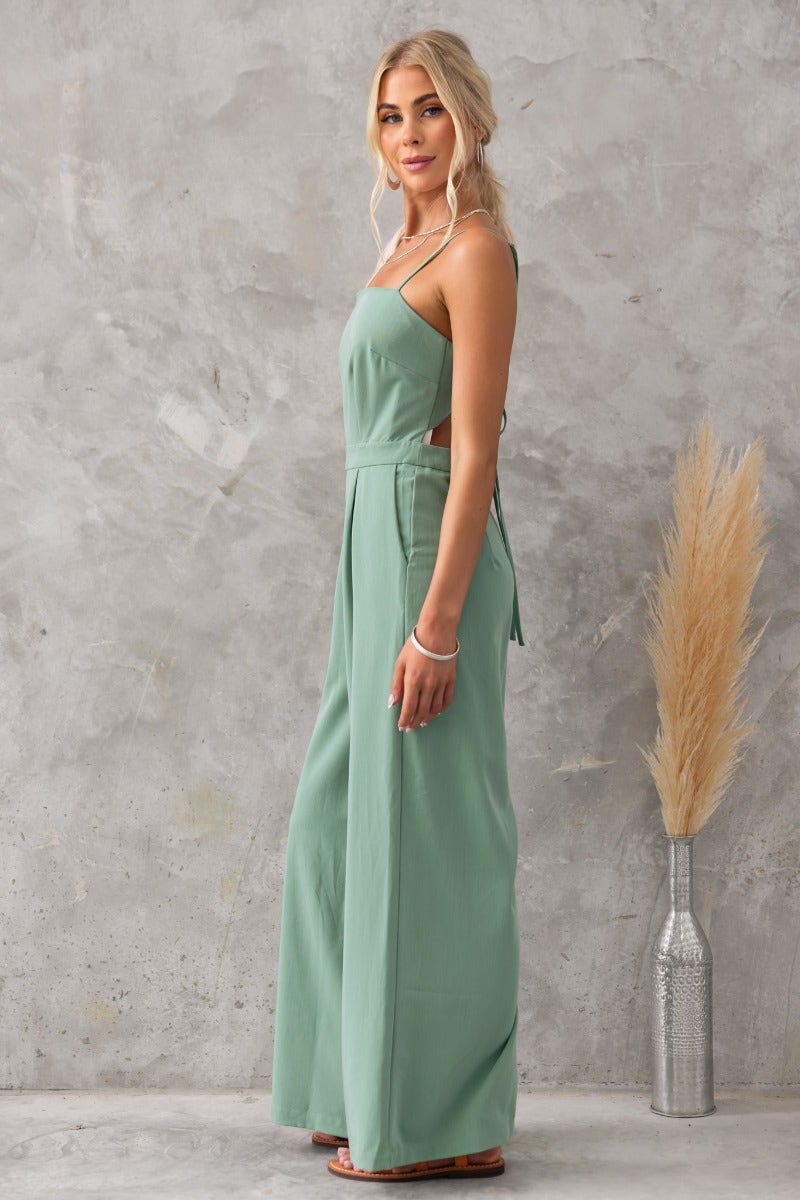 Model wearing the Greta Jumpsuit, a soft green jumpsuit with adjustable straps and a tie-back design.