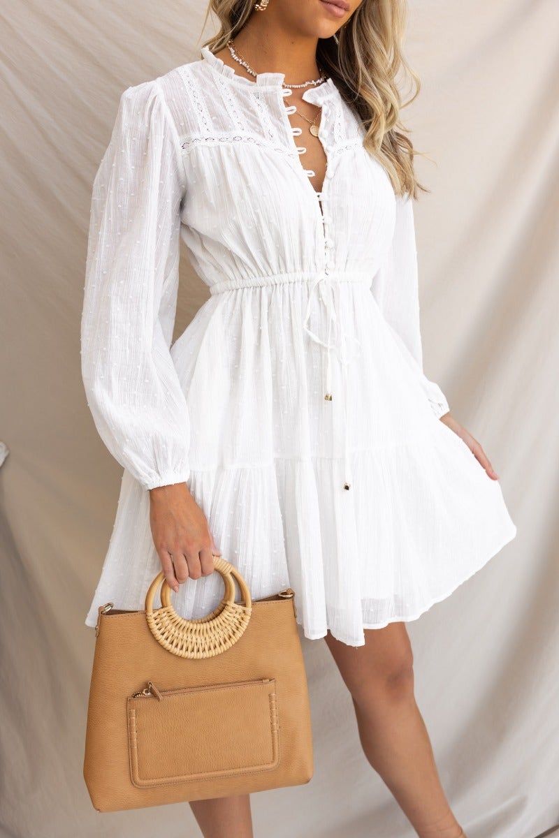 Model wearing a white cotton Alita dress with a button-up bust, elastic waistband, and pockets, holding a tan handbag.