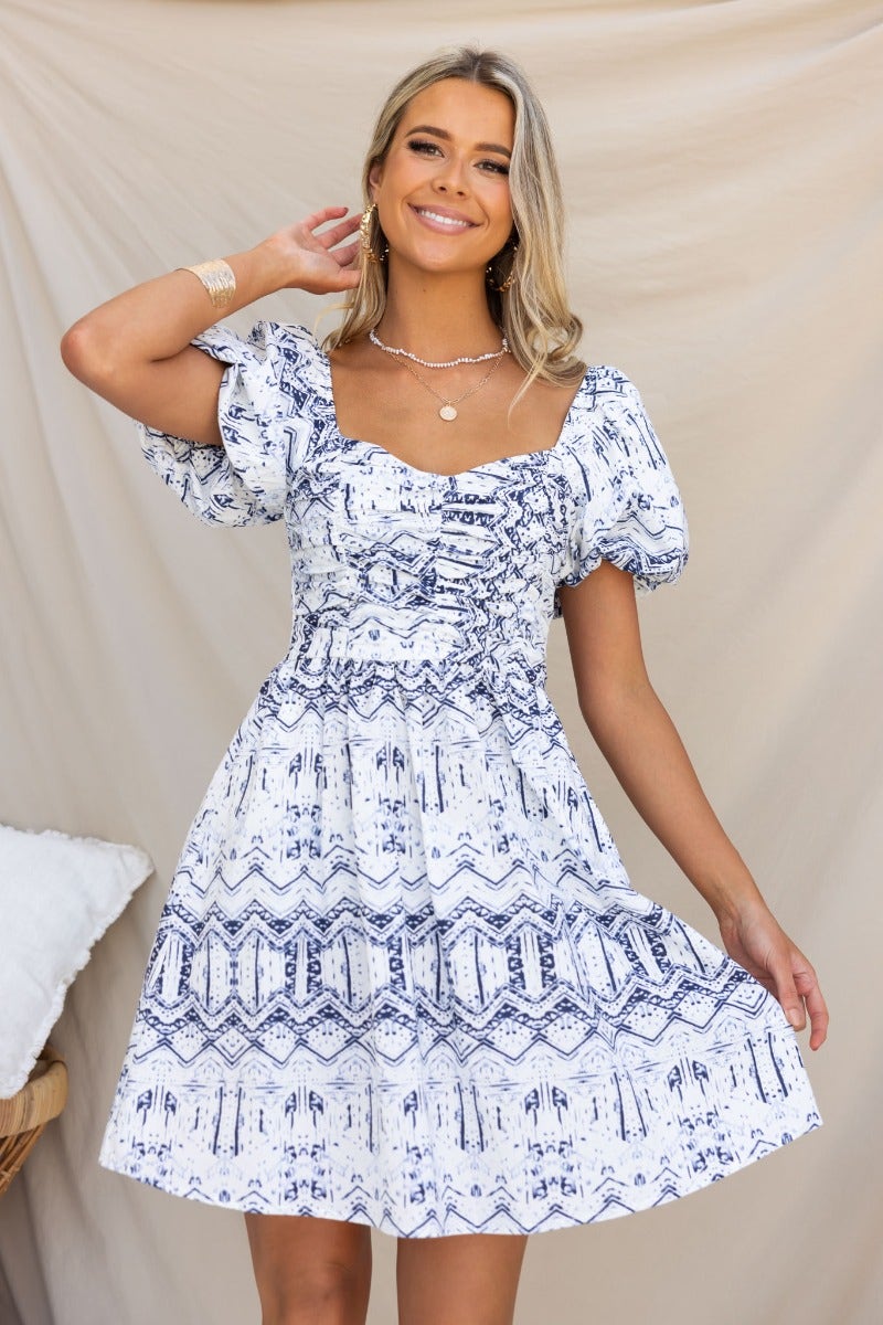 Model wearing a white and blue patterned cotton Harvie Dress with puff sleeves and a fitted bodice.