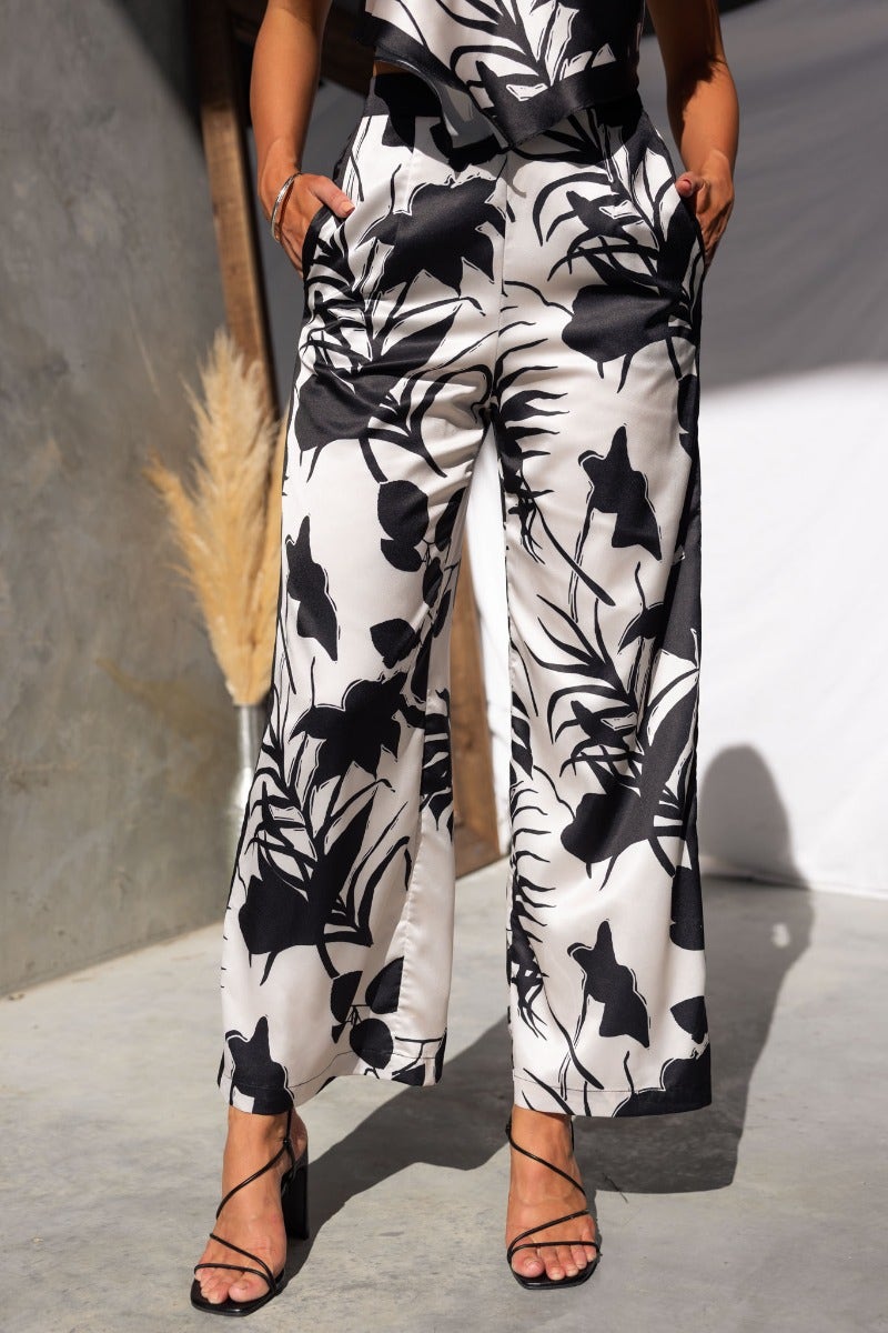 Persia Pants: Black and white satin pants featuring leafy prints, elastic waist, and pockets, styled for a chic summer look.
