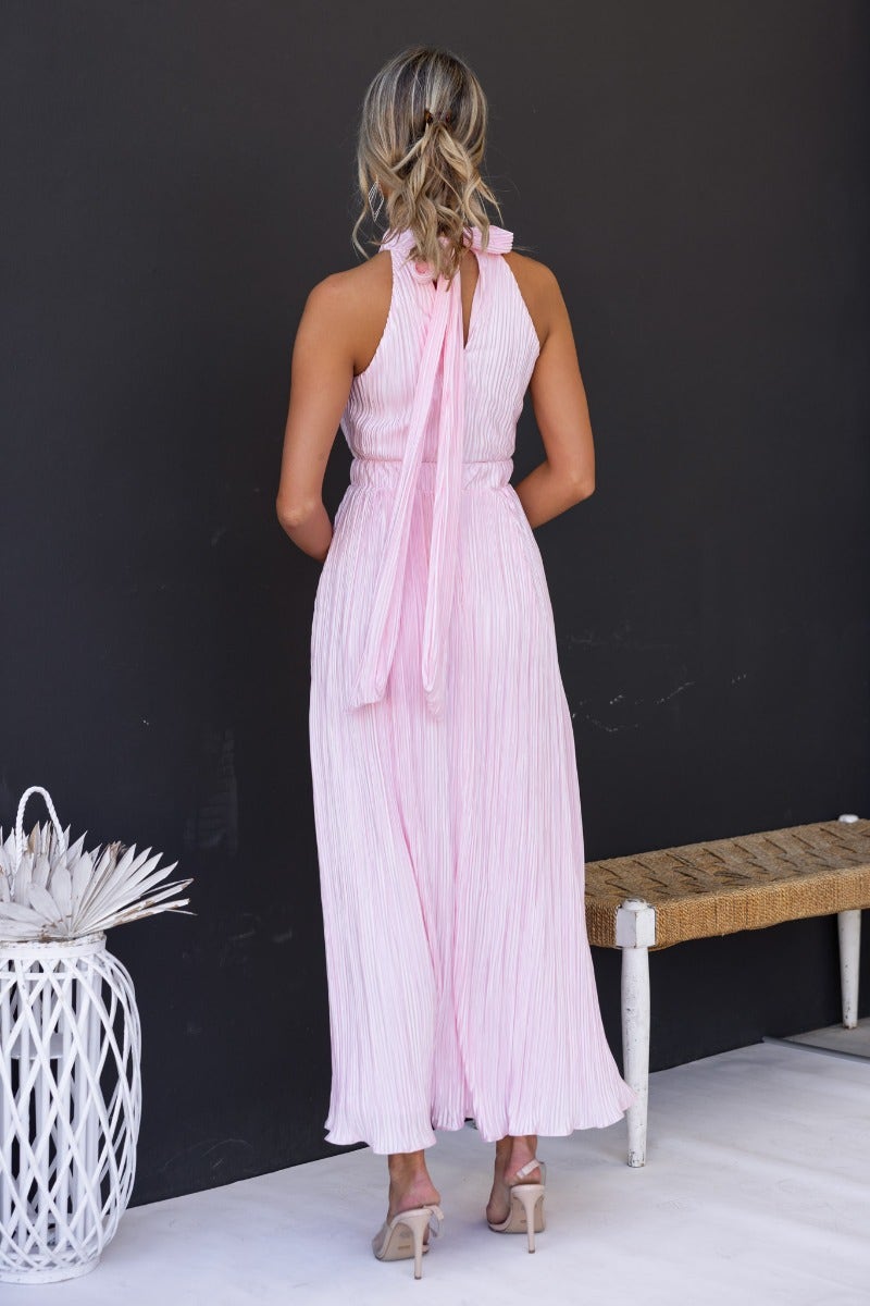 Back view of the Tyran Maxi Dress in pink with accordion pleats and a high tie neckline, styled elegantly.