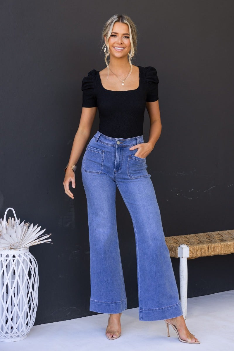 Model wearing a black textured bodysuit with cap sleeves and blue wide-leg jeans, posing against a dark backdrop.