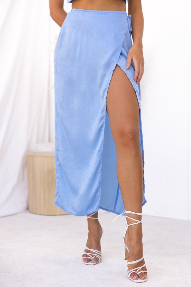 Nerida Wrap Midi Skirt in light blue with a high slit, styled with strappy heels, showcasing its asymmetric hem.