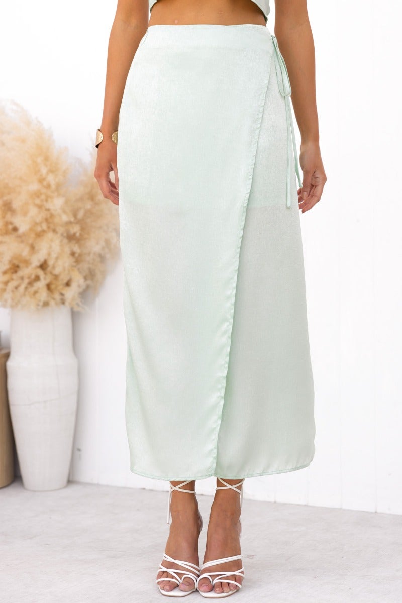 Nerida Wrap Midi Skirt in mint green, featuring a side tie and asymmetric hem, styled with strappy sandals.
