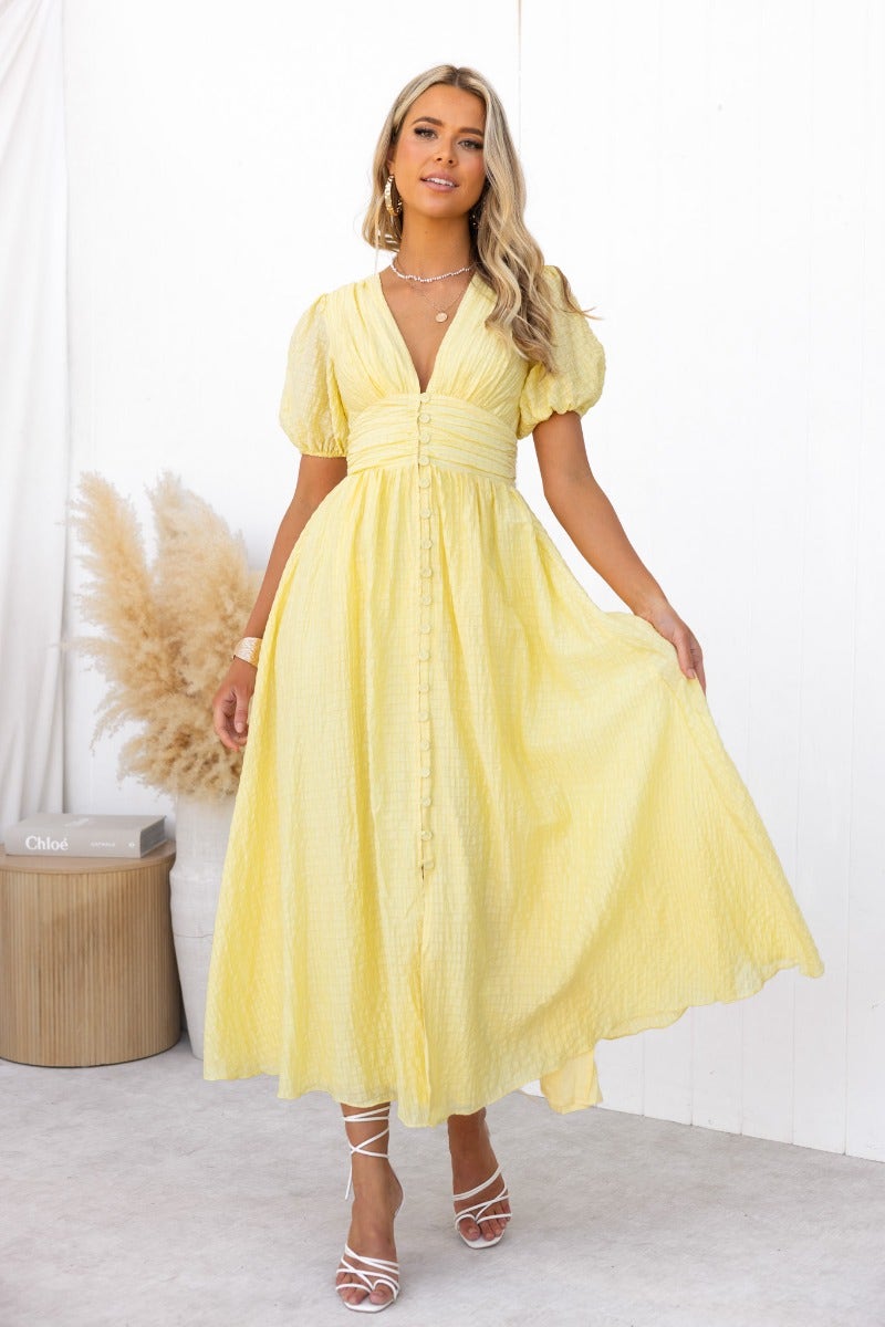 Model wearing a yellow textured maxi dress with V-neckline and puff sleeves, showcasing a stylish faux button-up front.