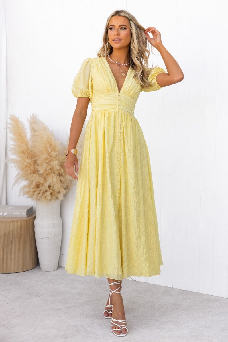 Hollie Maxi Dress in yellow with textured fabric, V-neckline, elastic sleeves, and faux buttons, styled in a bright setting.
