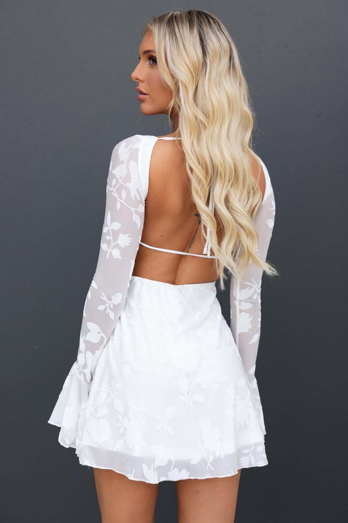 Back view of a model wearing a white Catiana Dress with floral design, long sleeves, and a tie back.
