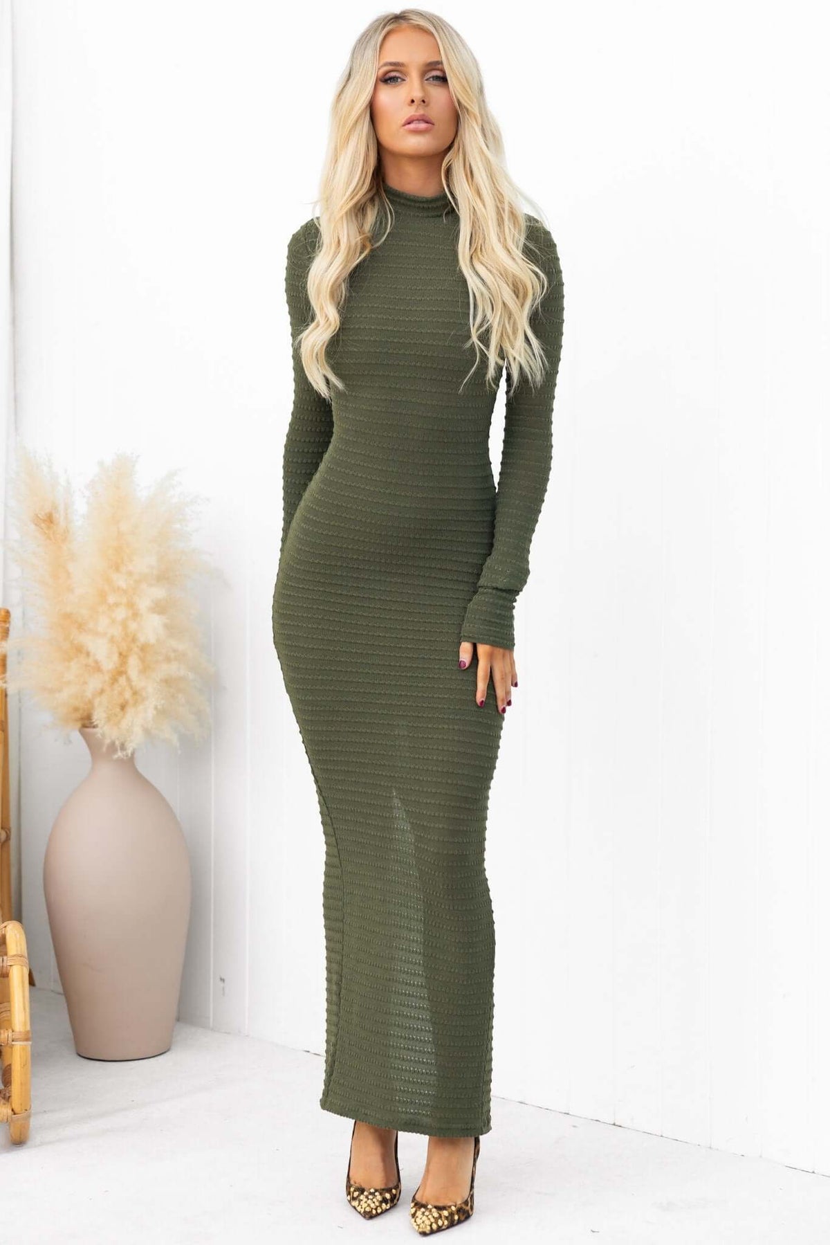 Everlie maxi dress in olive green with high neckline, long sleeves, and back split, styled with heels and a vase backdrop.