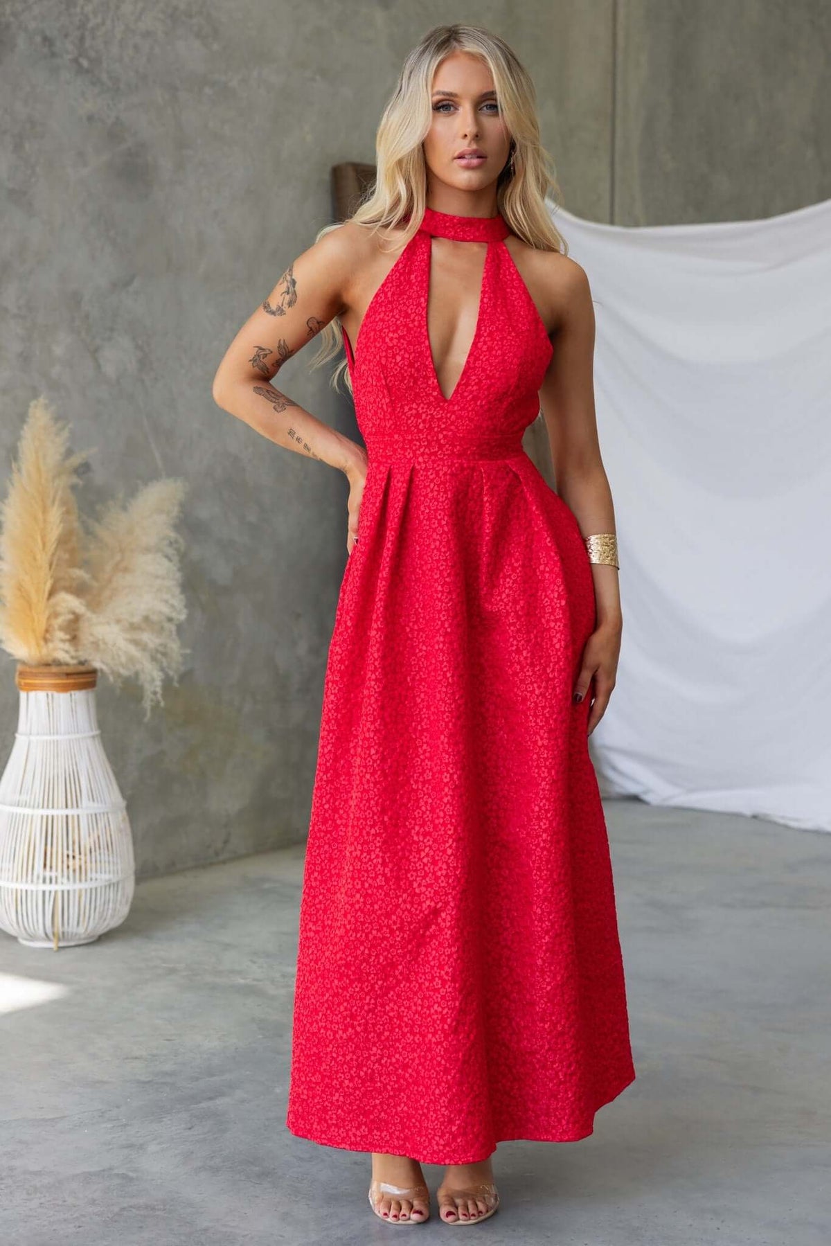 Model wearing a red Ultima Maxi Dress with halter neck, pleated waist, and textured detail against a minimalist background.