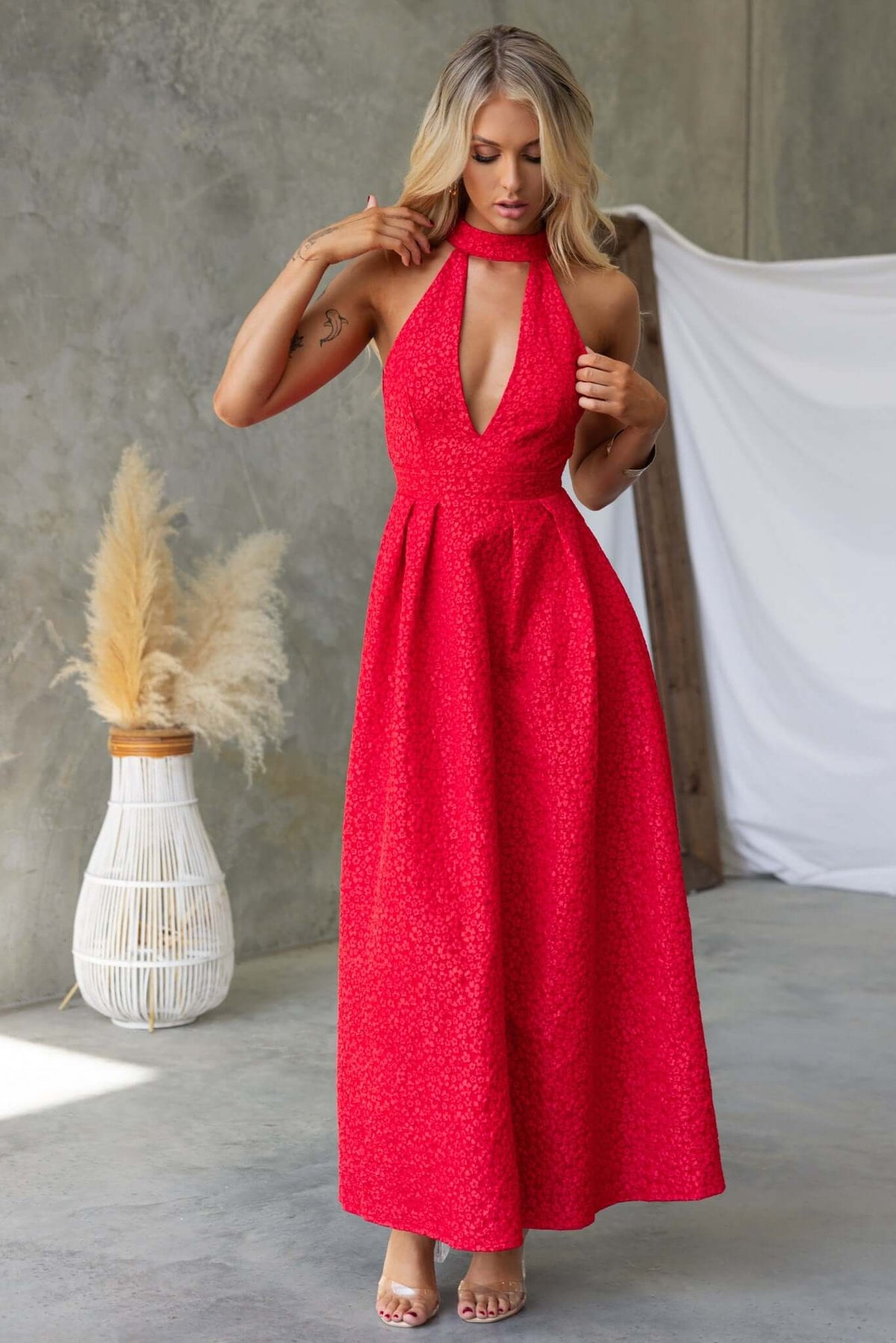 Stylish woman wearing a red Ultima Maxi Dress with a halter neck and pleated waist in a chic indoor setting.