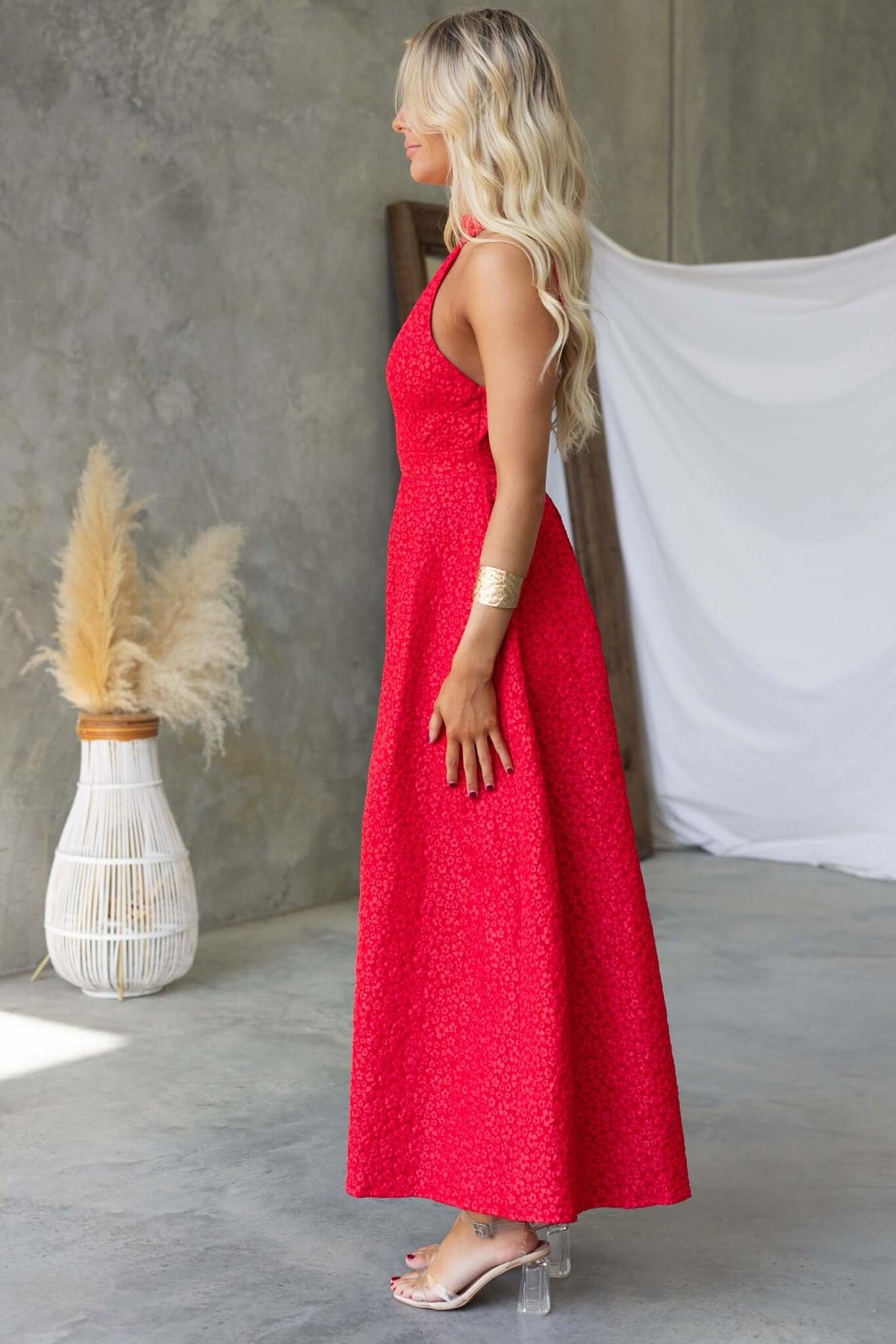 Model wearing a vibrant red Ultima Maxi Dress featuring a pleated waist and elegant design in a modern setting.
