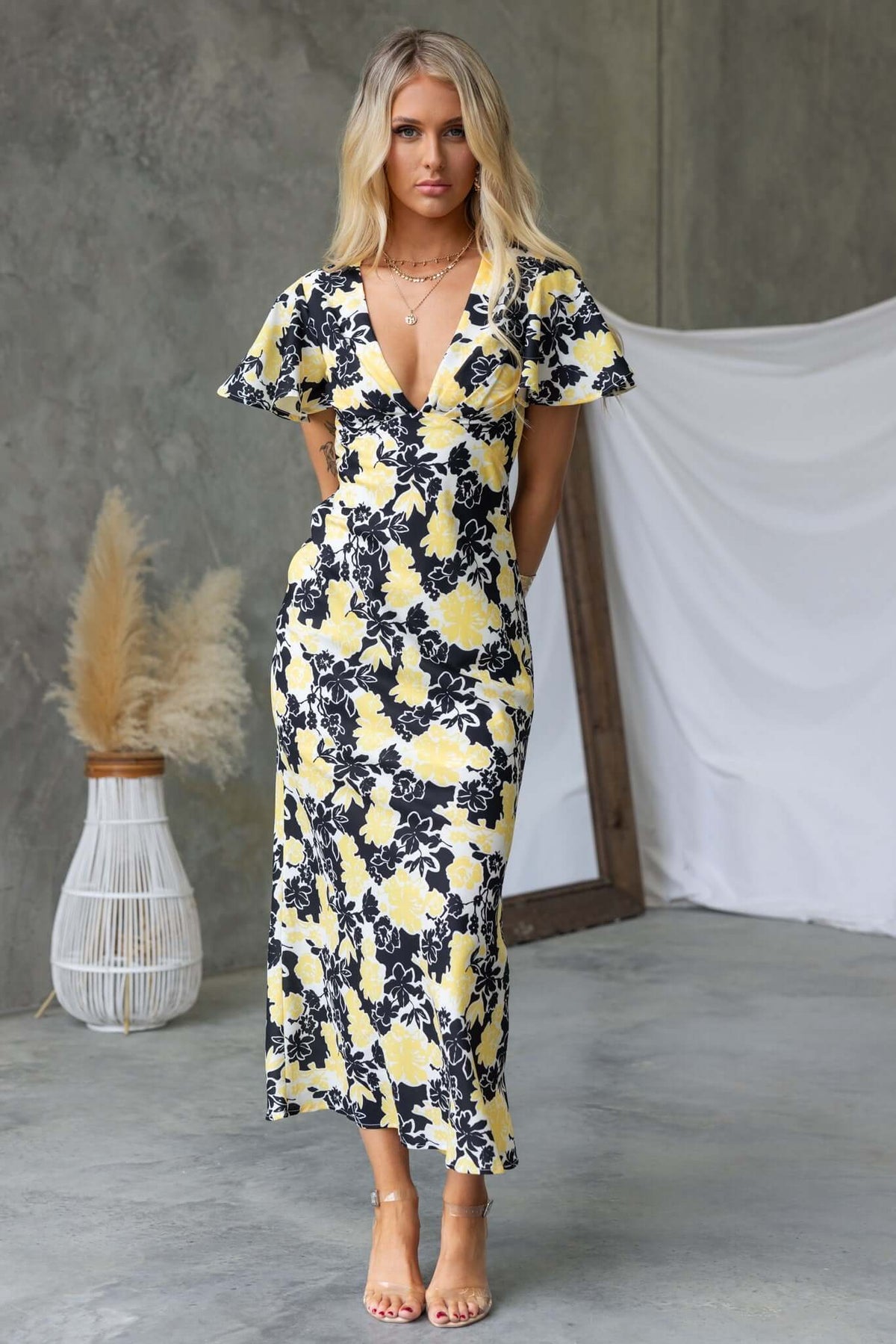 Model wearing a floral black and yellow Bartow Maxi Dress with V-neckline and short sleeves, styled in a modern indoor setting.