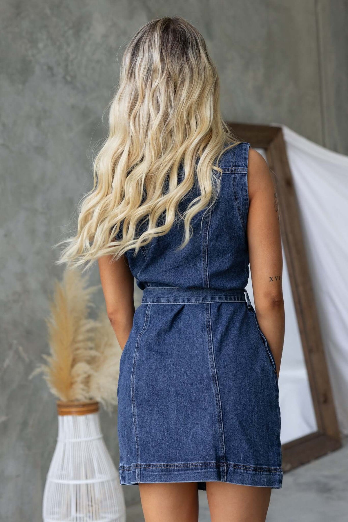 Back view of a woman in a Pokey Denim Dress, featuring a belted waist and pockets against a textured background.