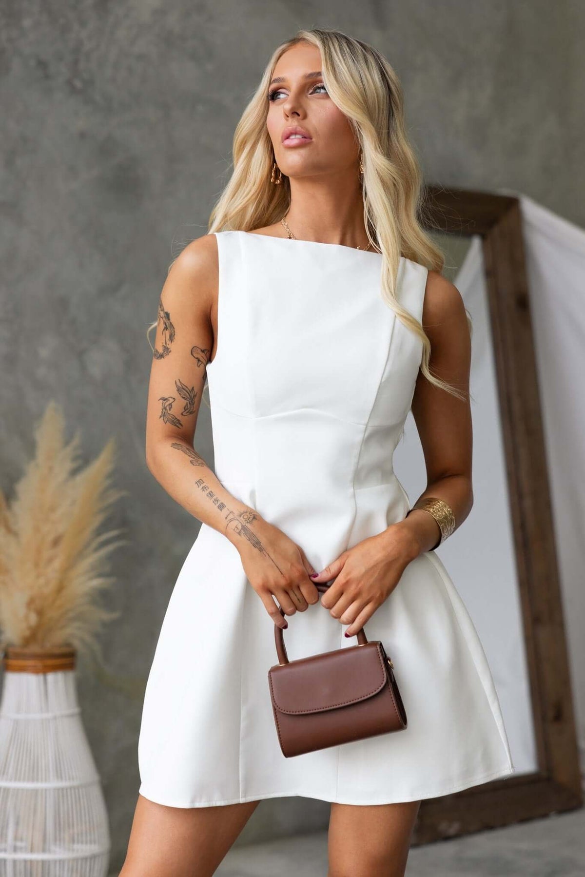 Model wearing a stylish white demure dress with a scoop back and holding a brown handbag.