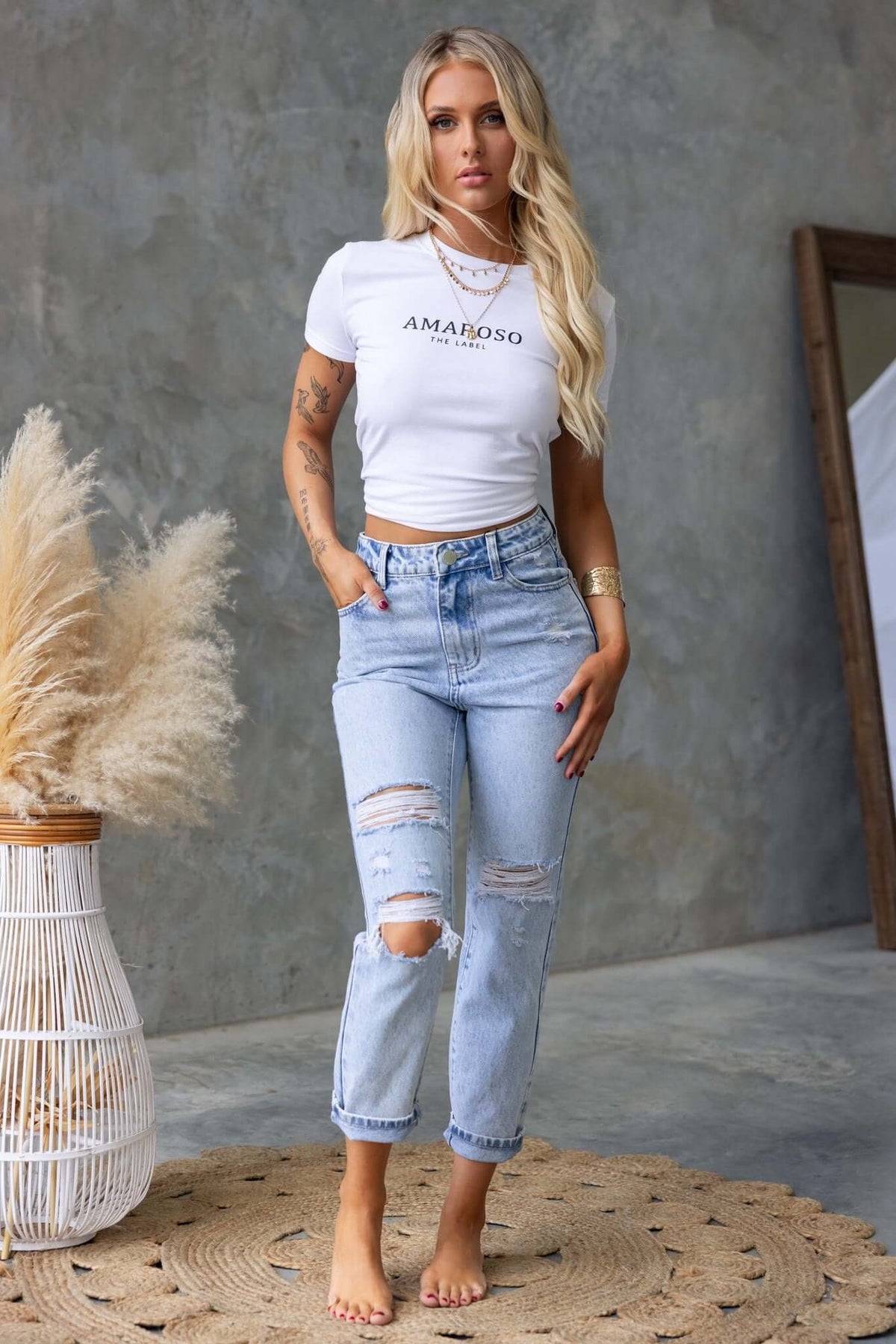 Model wearing Niles Jeans, mid-high waist denim with distressed details, styled with a white tee and beachy decor.