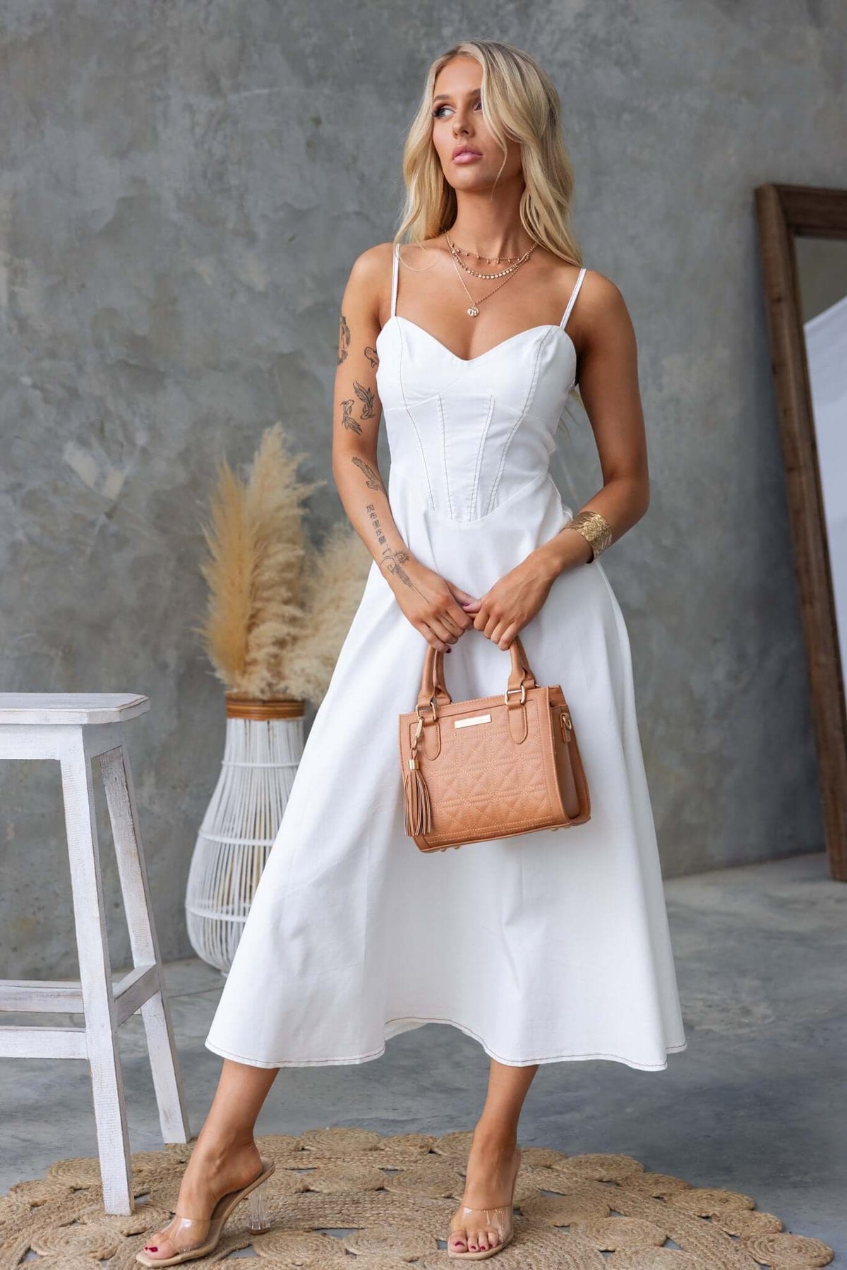 Stylish woman in a white midi dress with adjustable straps, paired with a tan handbag, set against a modern backdrop.