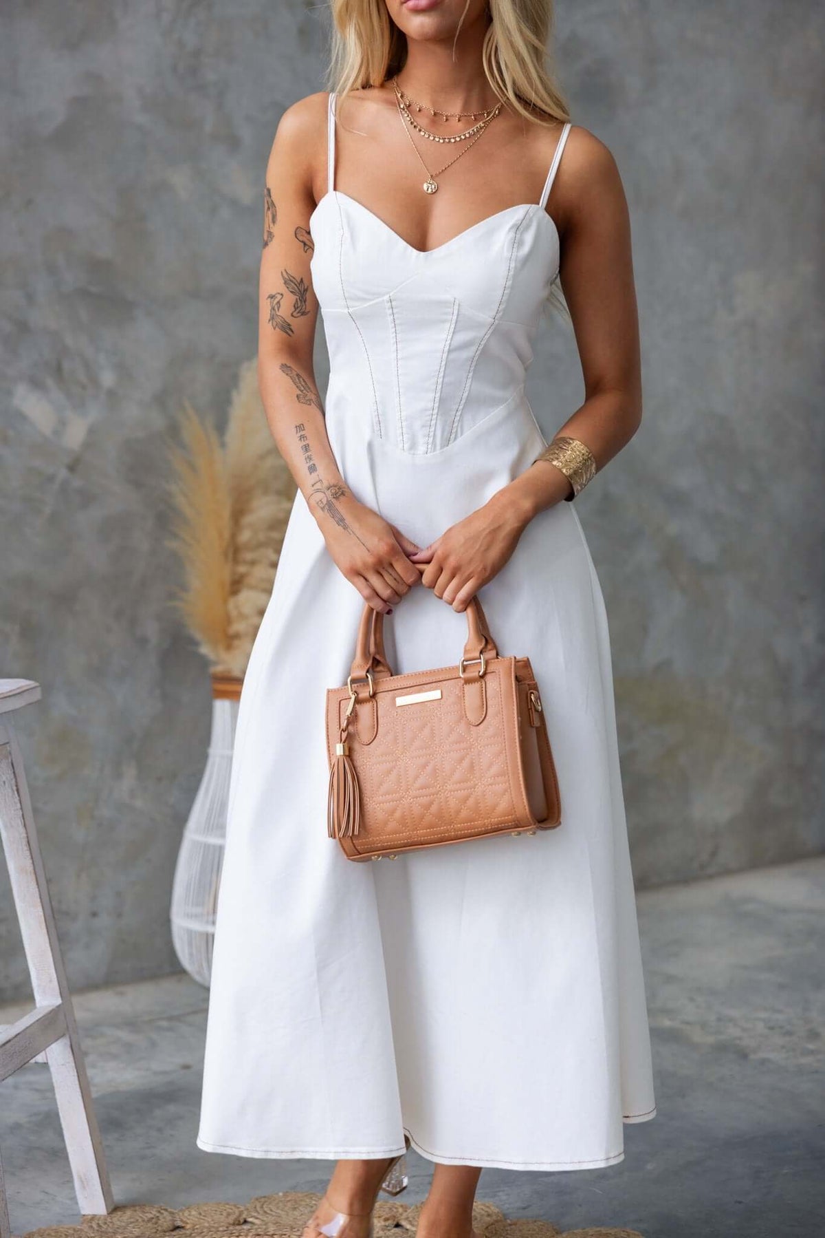 Model wearing a white denim midi dress with adjustable straps, holding a tan handbag, standing against a textured background.