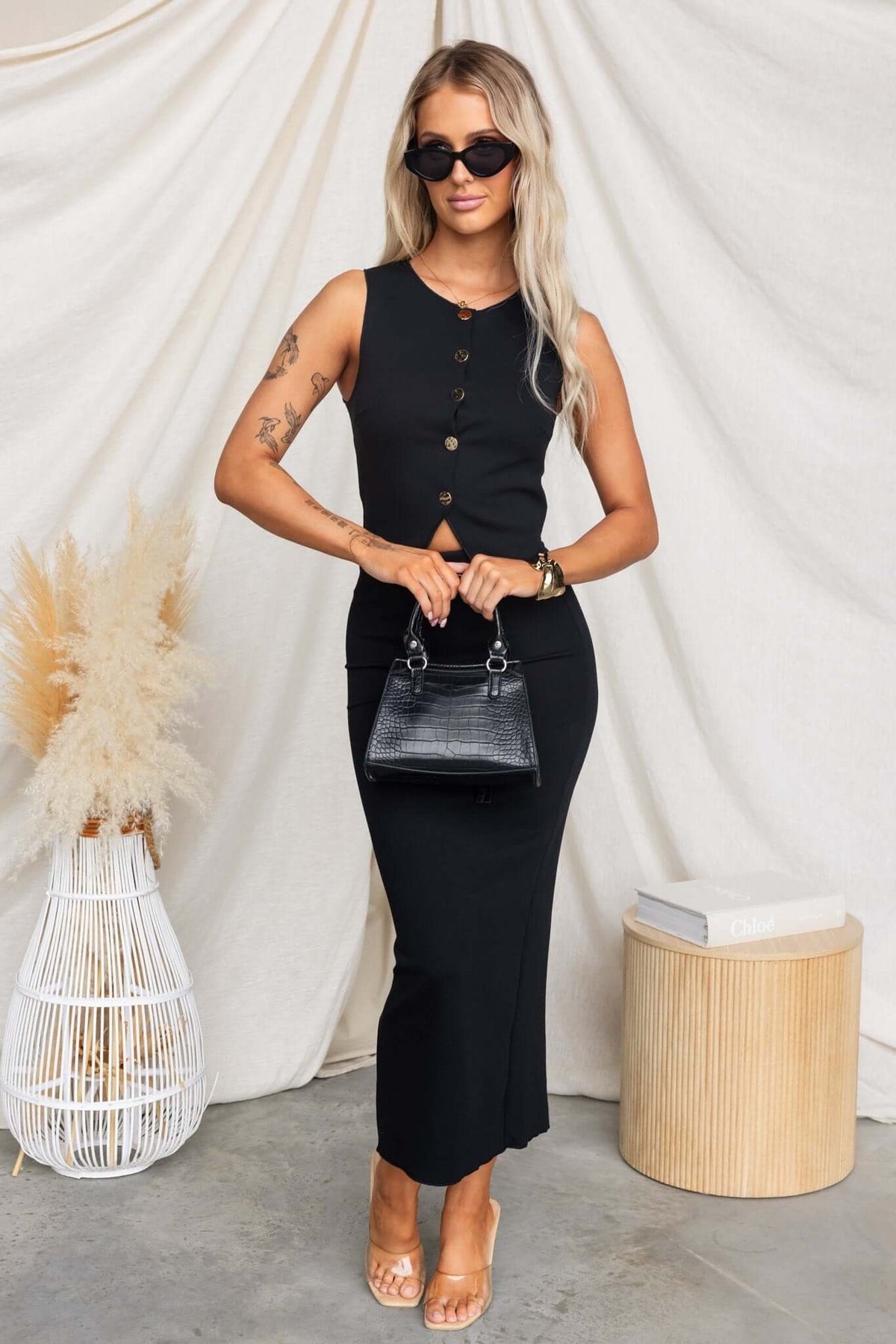 Stylish woman wearing Summit Midi Skirt with button details, sunglasses, and a handbag, set against a neutral backdrop.