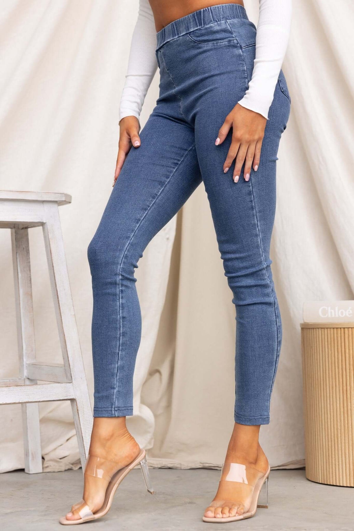 Model wearing Durant Jeans, mid-high waist denim jeggings with elastic waistband and back pockets, styled with clear heels.