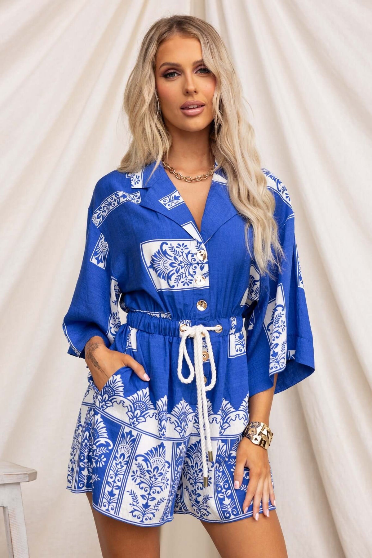 Blue and white floral Fiddler playsuit with collar, button-up front, elastic waistband, and pockets, modeled by a woman.