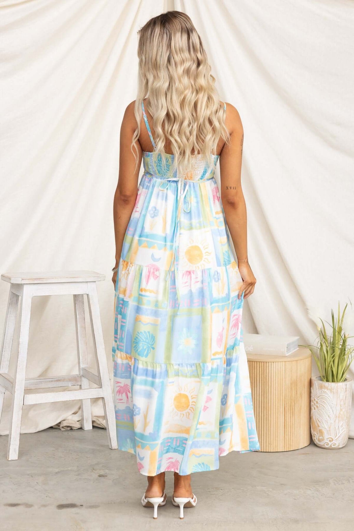 Back view of a woman in a colorful patterned maxi dress with adjustable straps and pockets, styled in a boho setting.