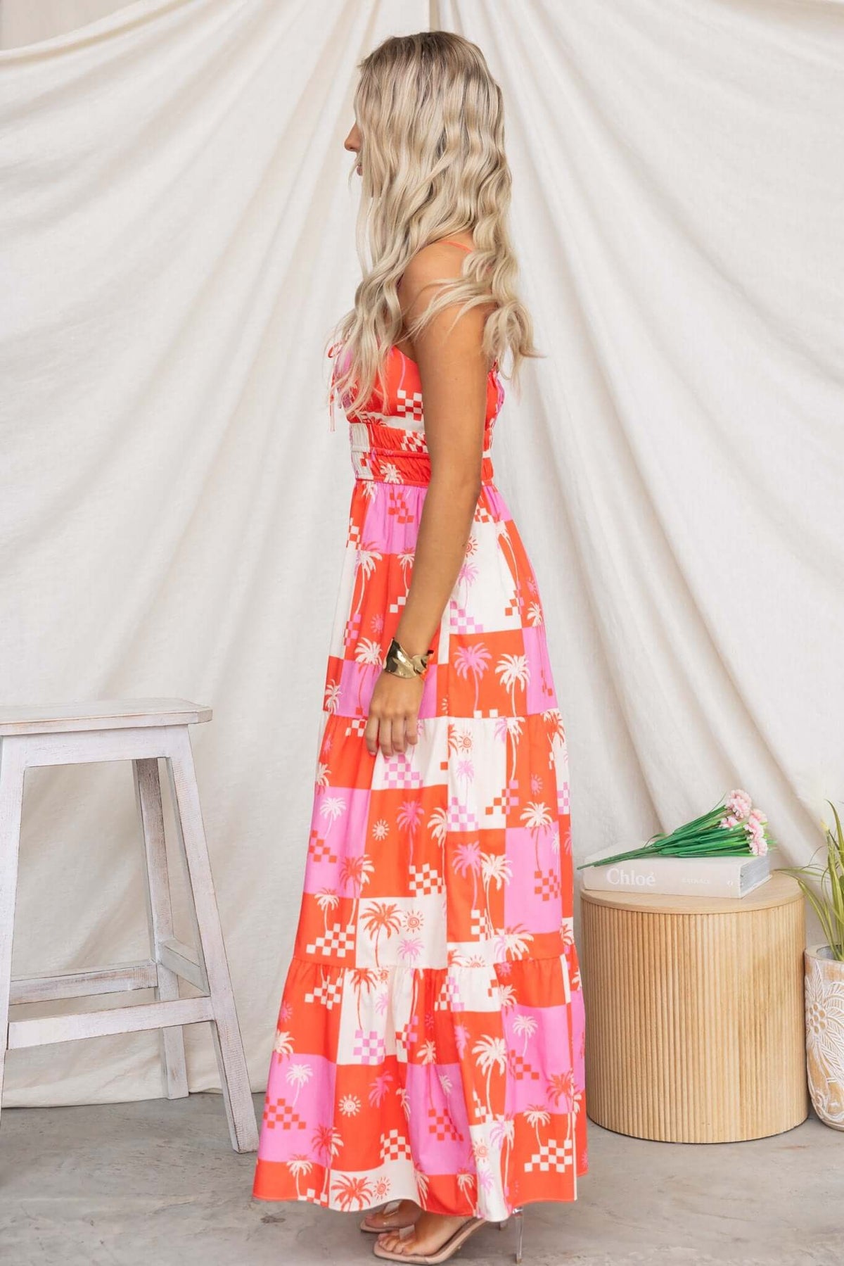 Side view of a woman in a Norment Maxi Dress, featuring vibrant orange and pink patterns, thin straps, and ruffled tiers.
