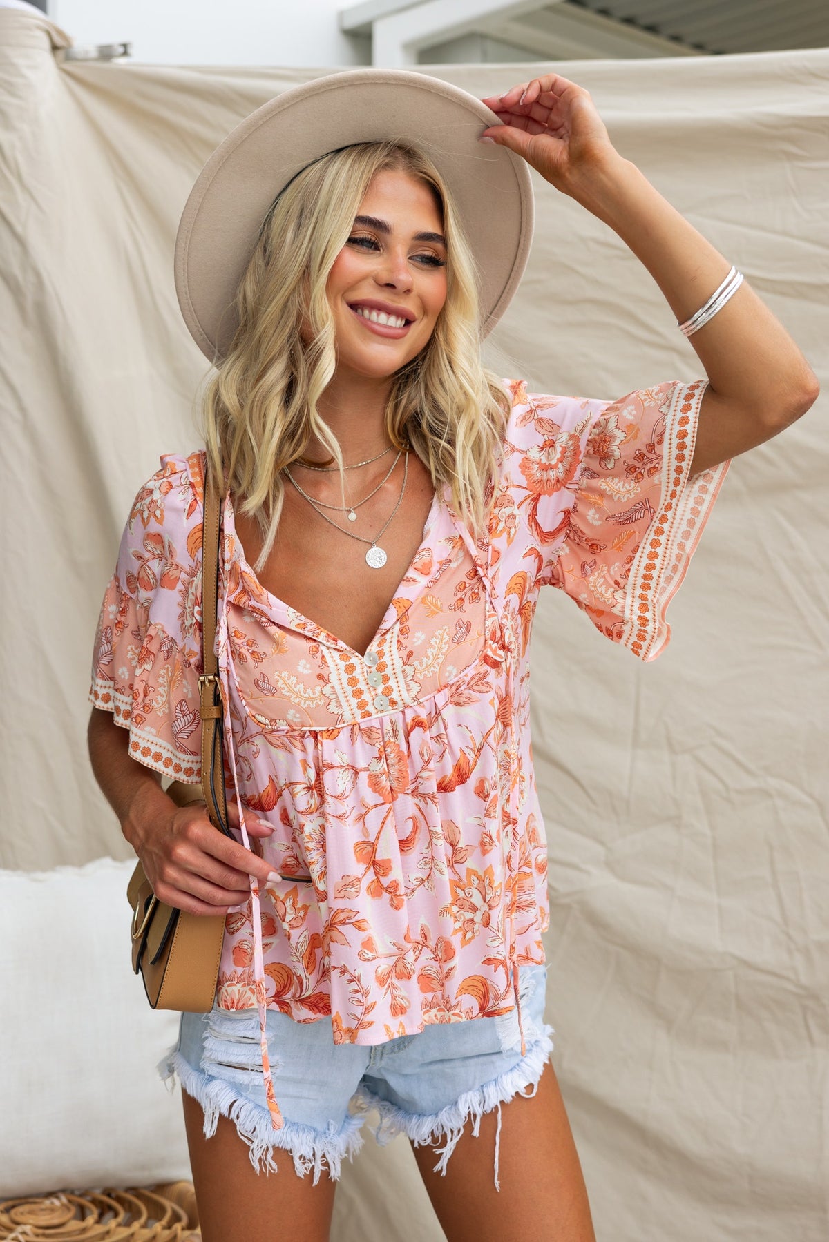 Smiling woman in floral print Reeva Top with V-neckline and hat, wearing denim shorts against a neutral backdrop.