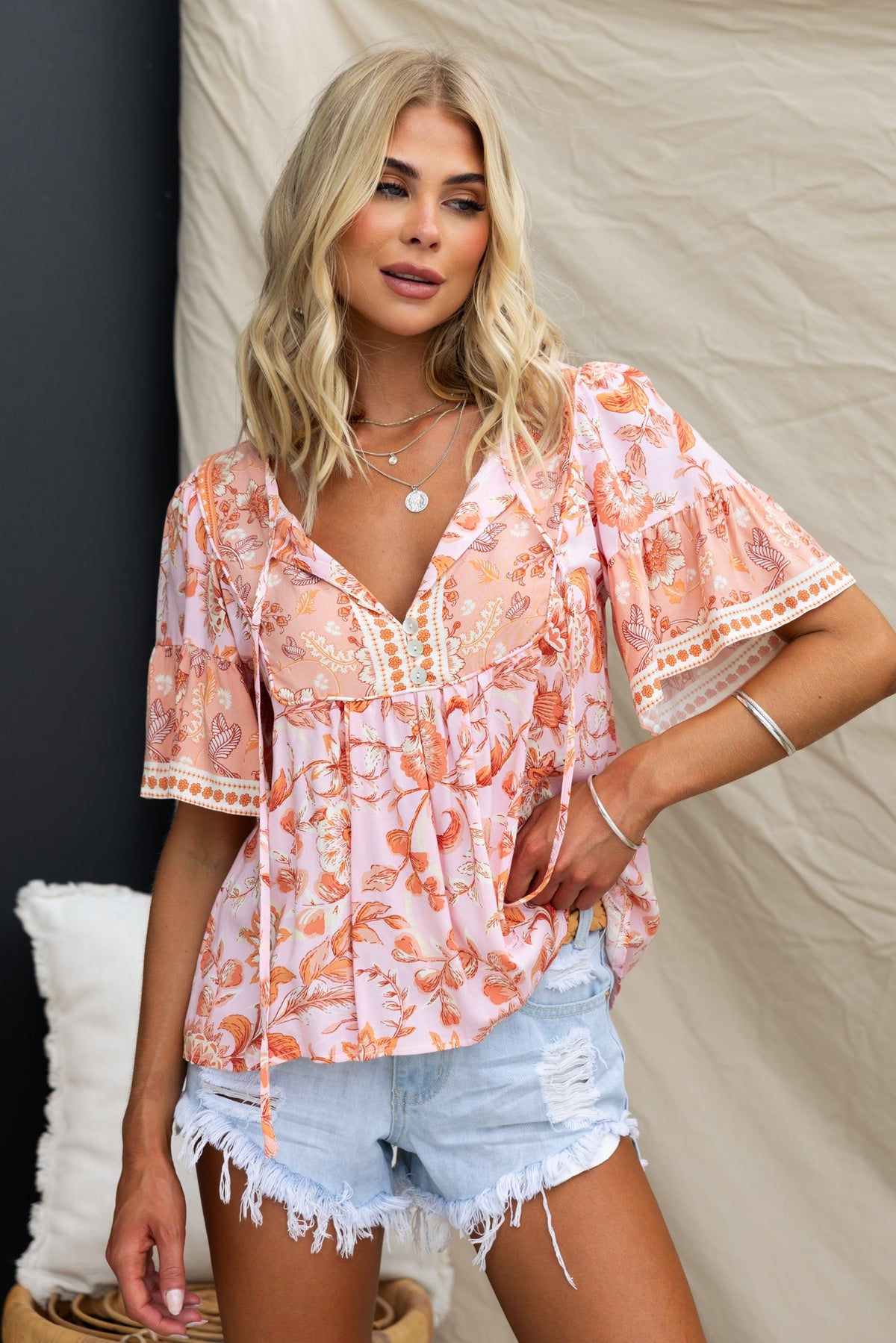 Model wearing a pink floral print Reeva Top with V-neckline and tie detail, styled with denim shorts.