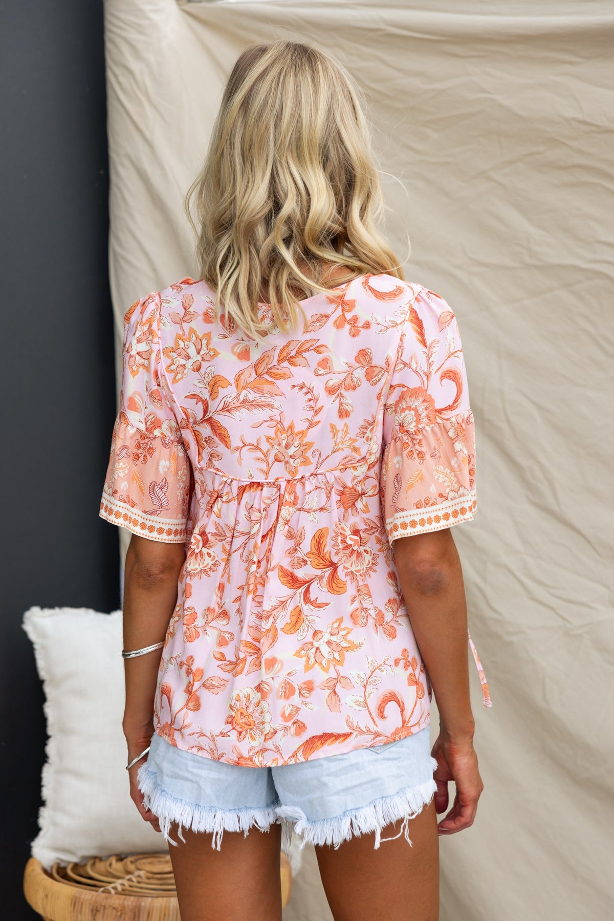 Back view of a woman wearing a floral print Reeva top with V-neck and tie detail, paired with denim shorts.