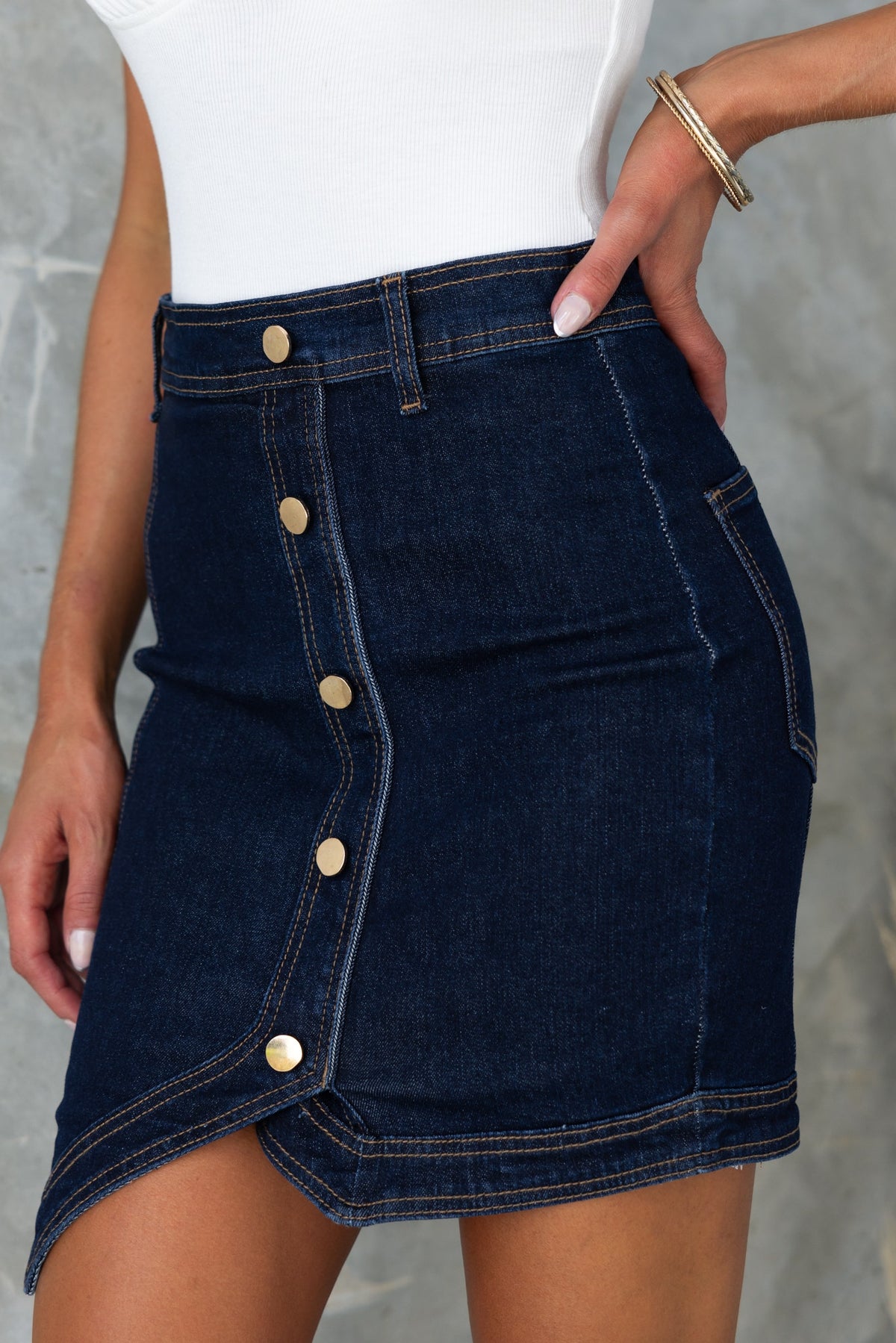 Dark navy denim skirt with faux buttons, mid-high waist fit, and lightly frayed hemline, styled with a white top.