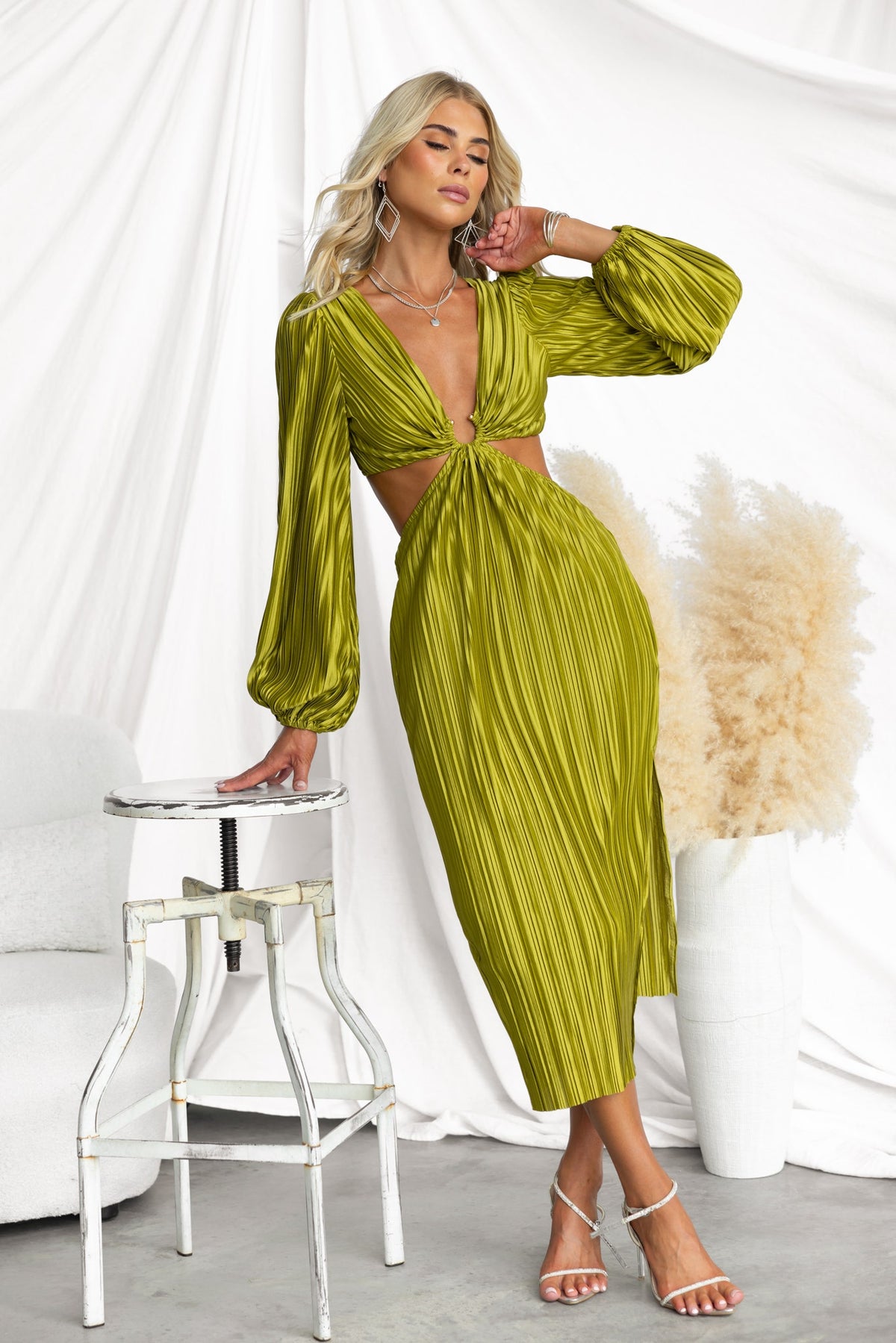Aulia Midi Dress in olive green with accordion pleats, V-neckline, and cut-out waist detail, styled with elegant accessories.