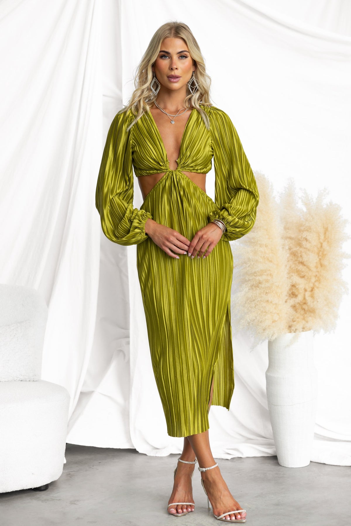 Aulia Midi Dress in olive green with pleats, V-neck, cut-out waist, and side split, styled with heels on a neutral backdrop.