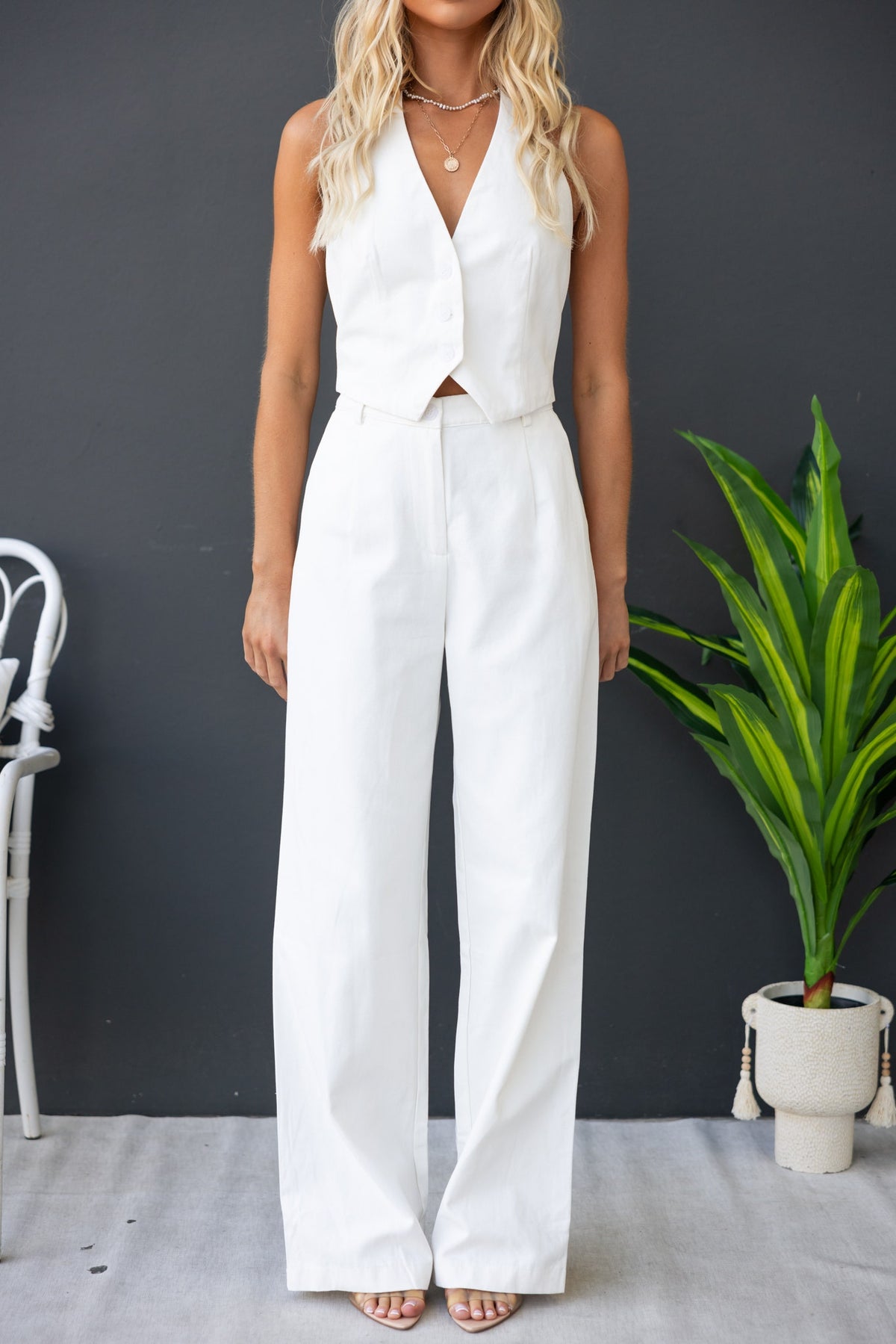 Model wearing a chic white vest and wide-legged pants, posing against a dark background with greenery nearby.