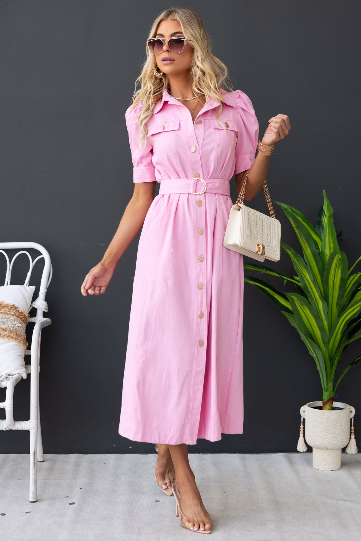 Model wearing a pink denim maxi dress with a collar, button-up front, removable belt, and styled with sunglasses.