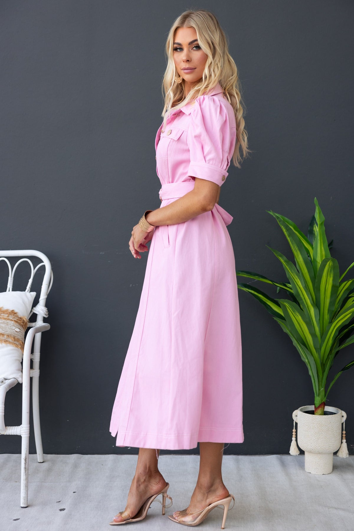 Model wearing a pink dress with puff sleeves and a tie waist, posed against a dark wall and green plant.