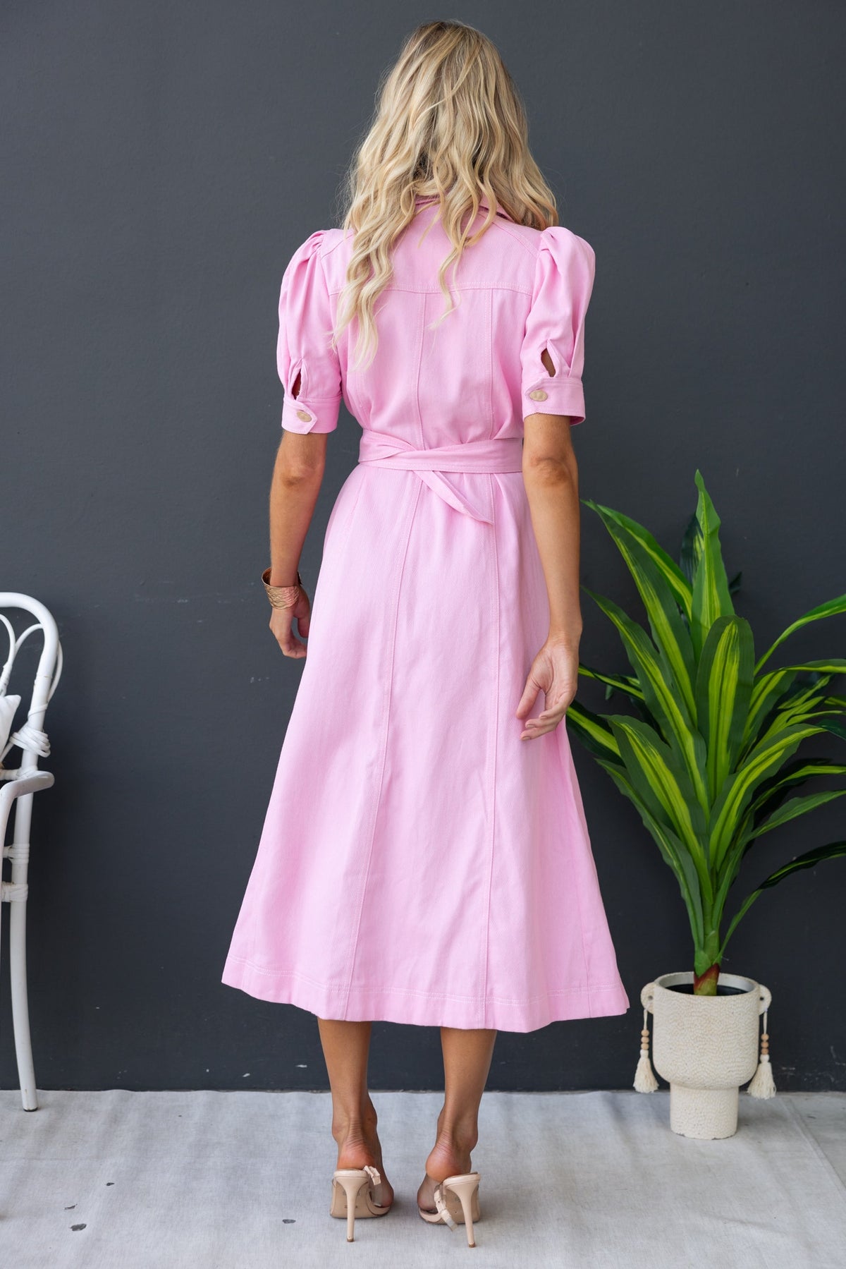 Back view of a pink denim maxi dress with puff sleeves and a waist tie, styled with a plant in a bright setting.