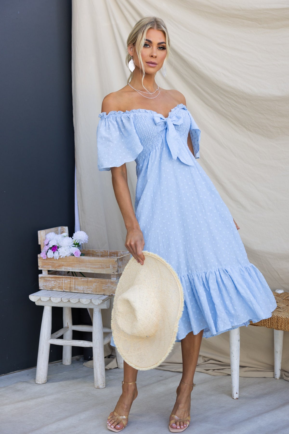 Angelis Midi Dress in light blue with off-shoulder neckline, tie at bust, and flutter sleeves, styled with a straw hat.