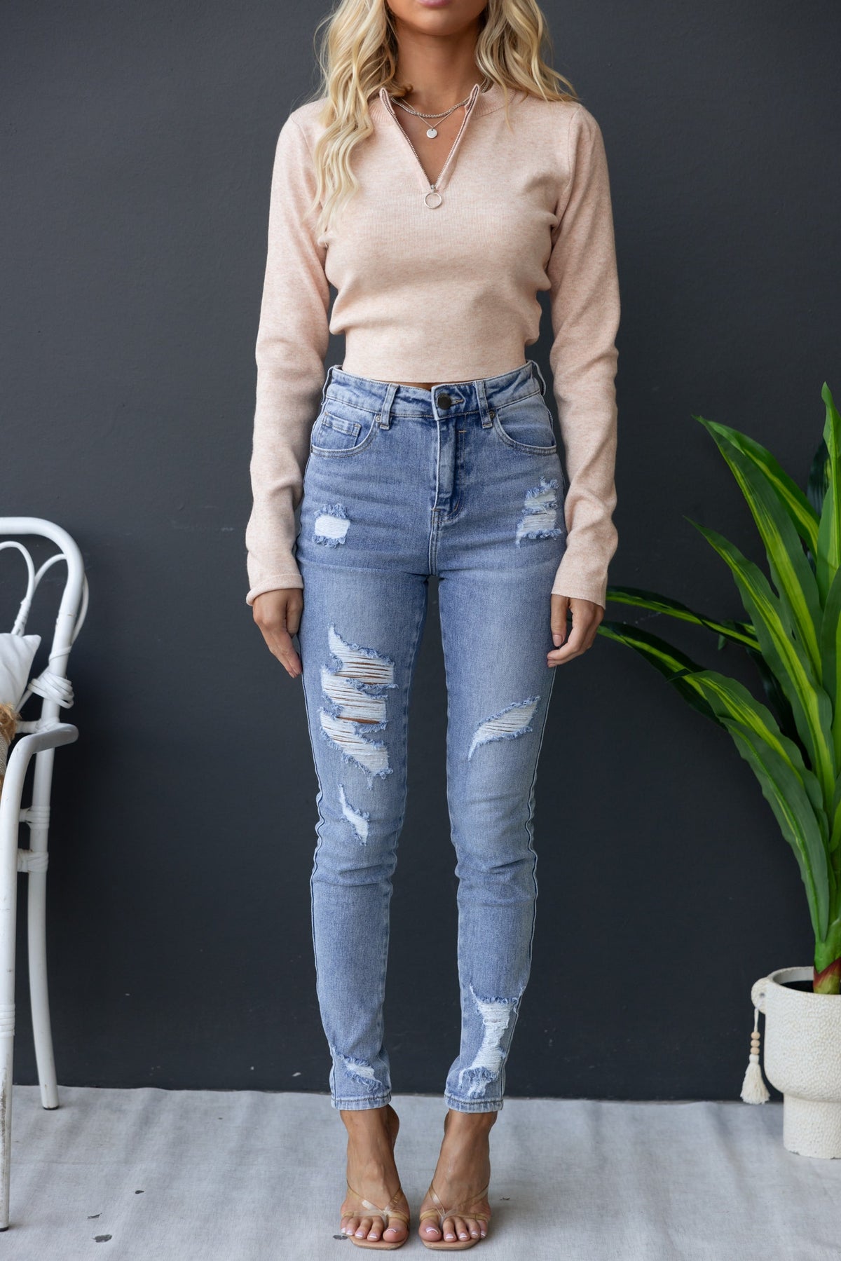 Woman wearing a beige high-neck zip-up Maura Top with distressed jeans and sandals, styled with a minimalistic background.