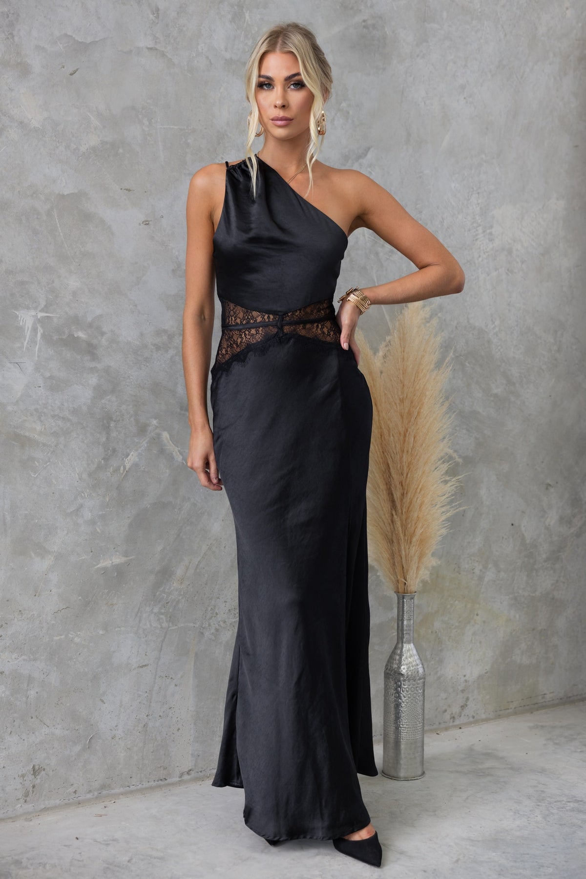 Model wearing a stylish black Bombshell Maxi Dress with lace detailing and a one-shoulder neckline, posed against a textured background.