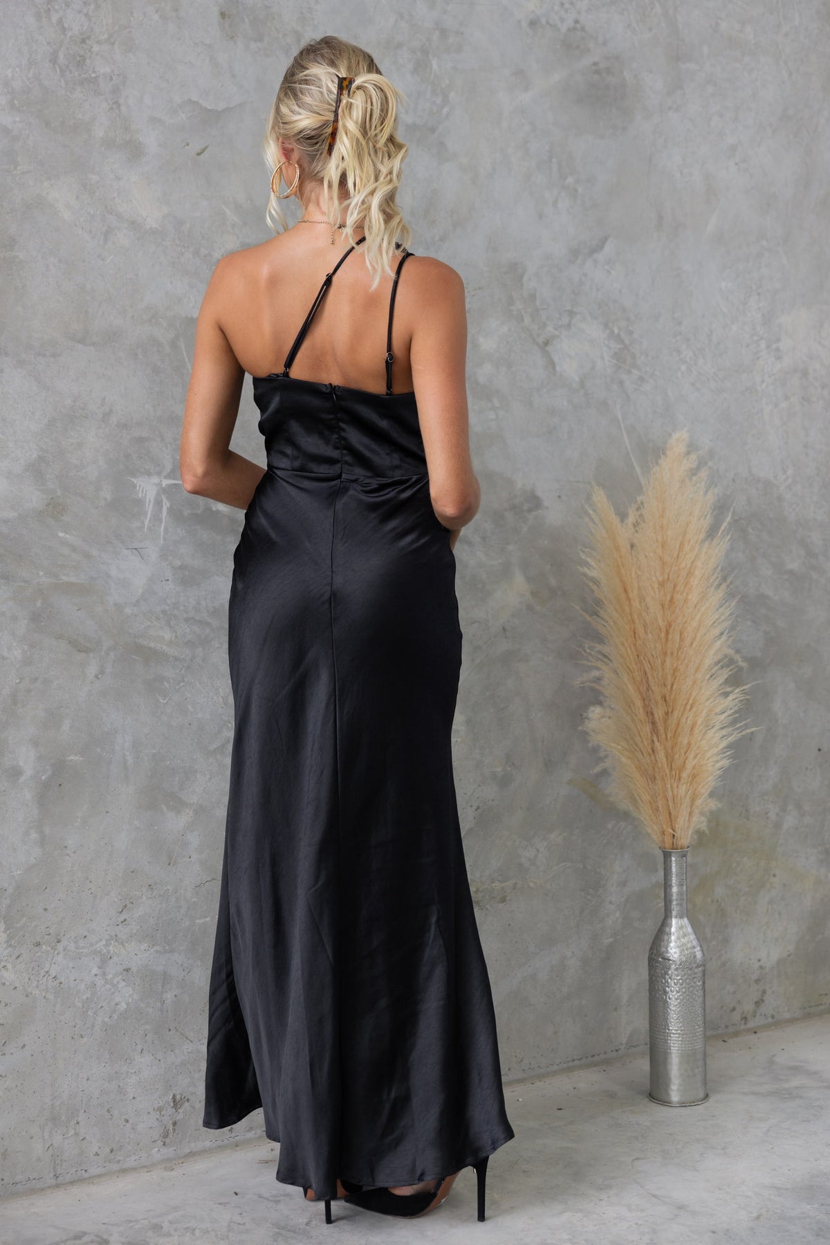 Back view of a woman in a black Bombshell Maxi Dress with one-shoulder neckline and adjustable straps.