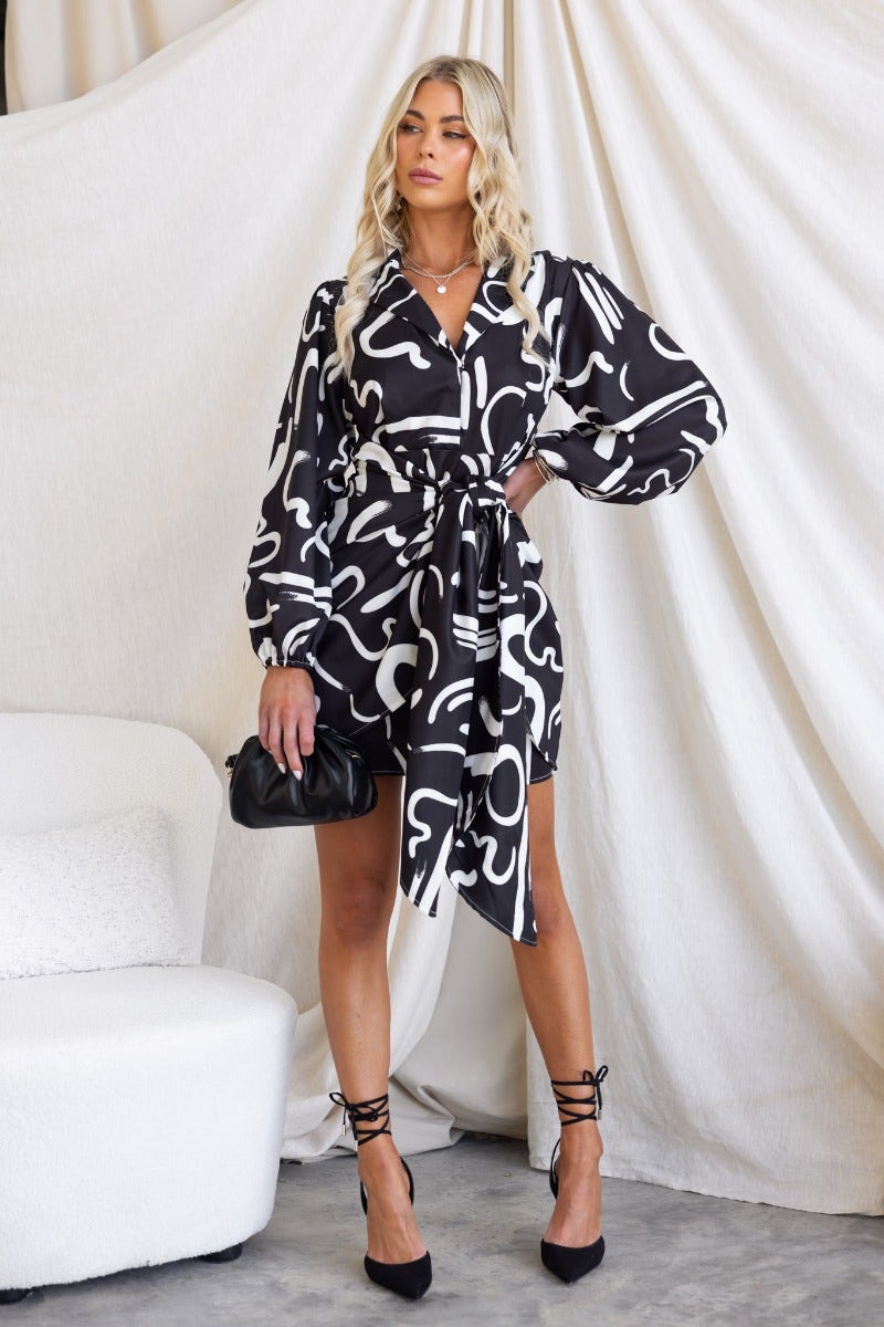 Yuliana Dress in black print with V-neck, button detail and wrap tie, worn by a model in a stylish setting.