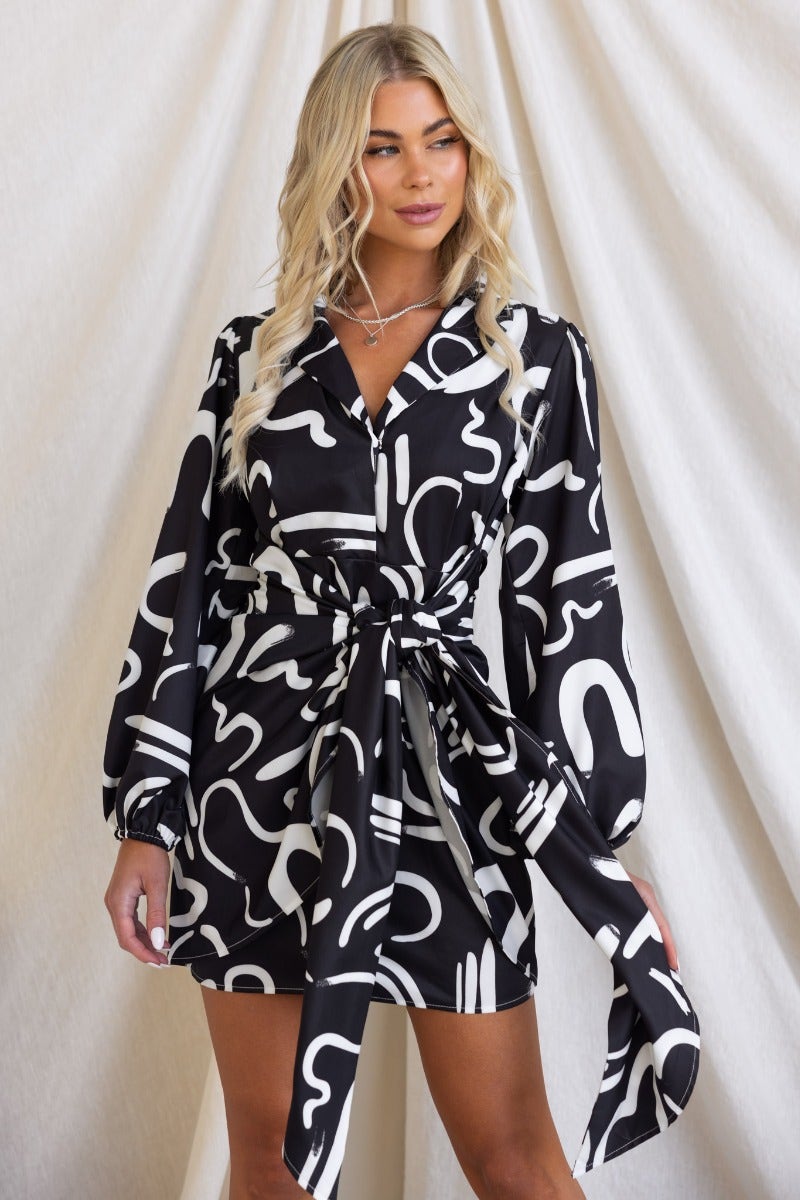 Model wearing the Yuliana Dress, a black print dress with a V-neckline and wrap tie detail.