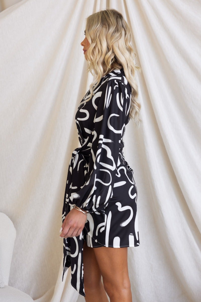 Yuliana Dress featuring a black and white abstract print, V-neck collar, and tie detail at the skirt, model shown side view.