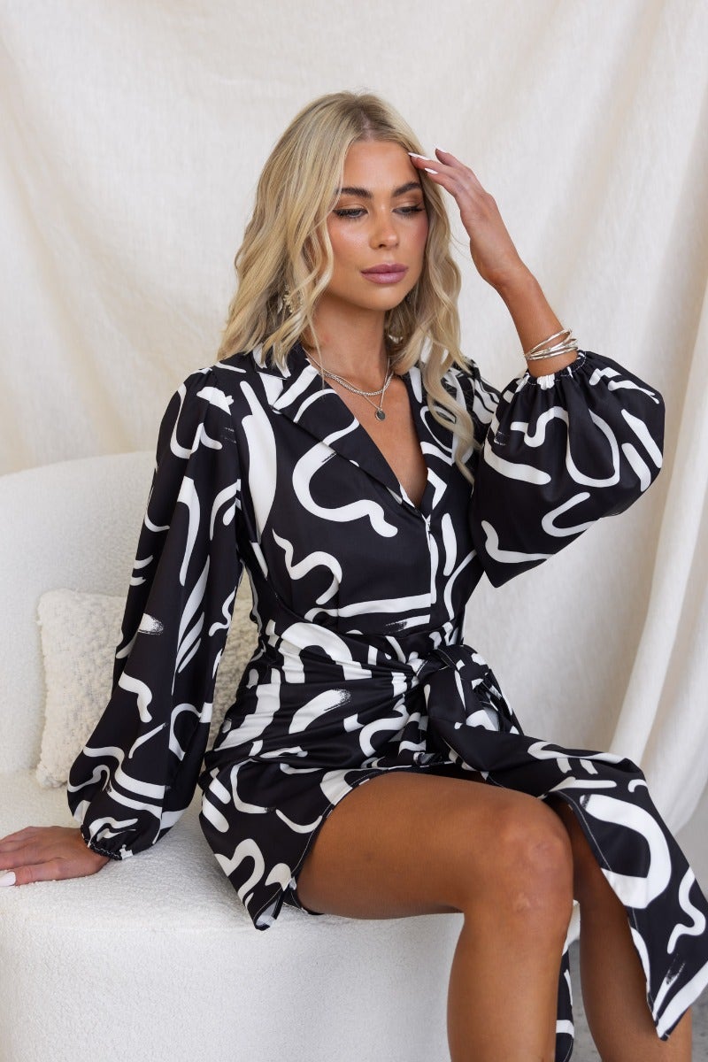 Model wearing Yuliana Dress, a black and white print with V-neck and wrap tie skirt, showcasing elegant style.